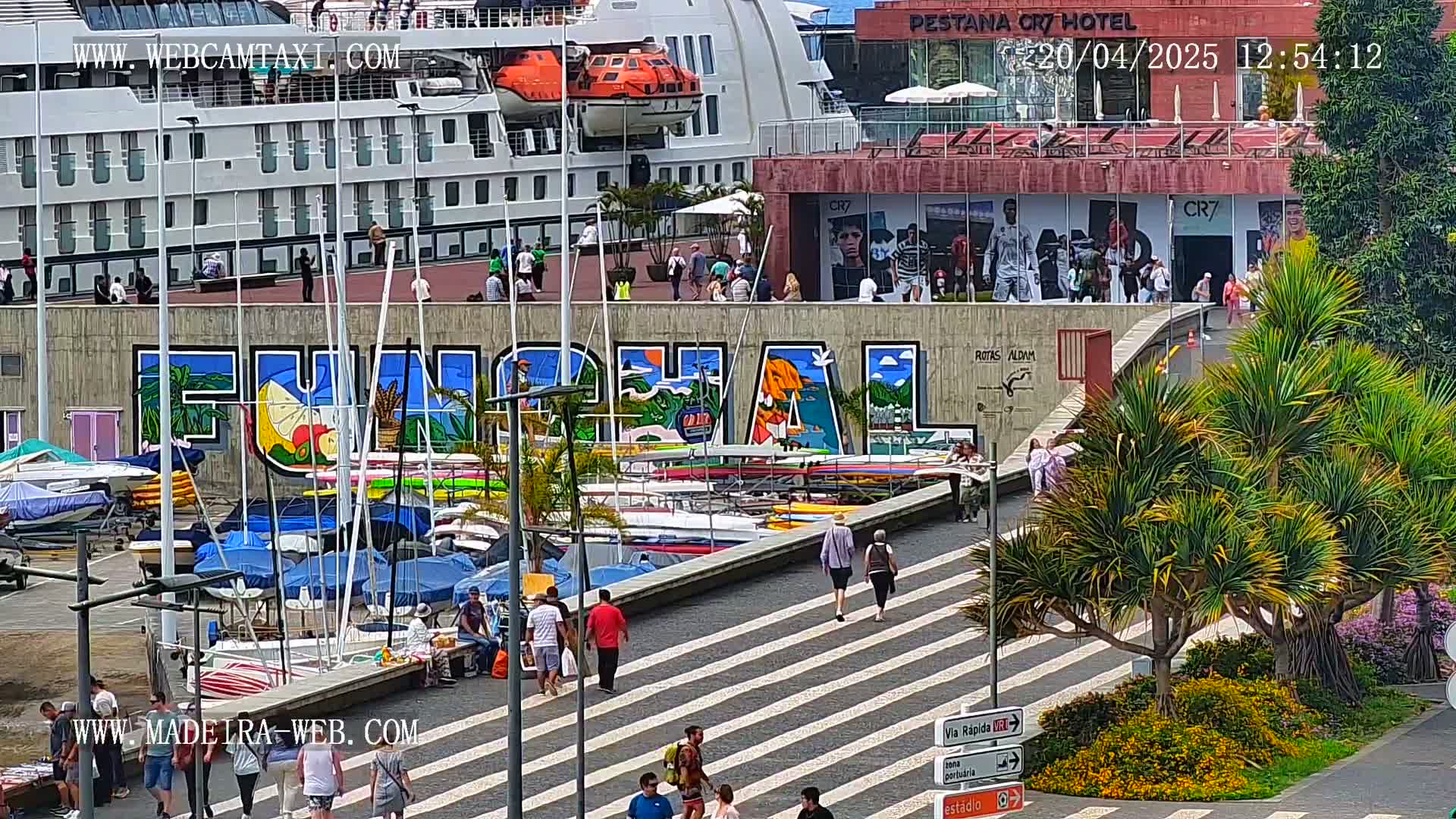 A sunny harbor scene shows a large cruise ship docked near a colorful mural of "Funchal," with numerous small boats moored below and people walking along a striped pedestrian walkway lined with palm trees.