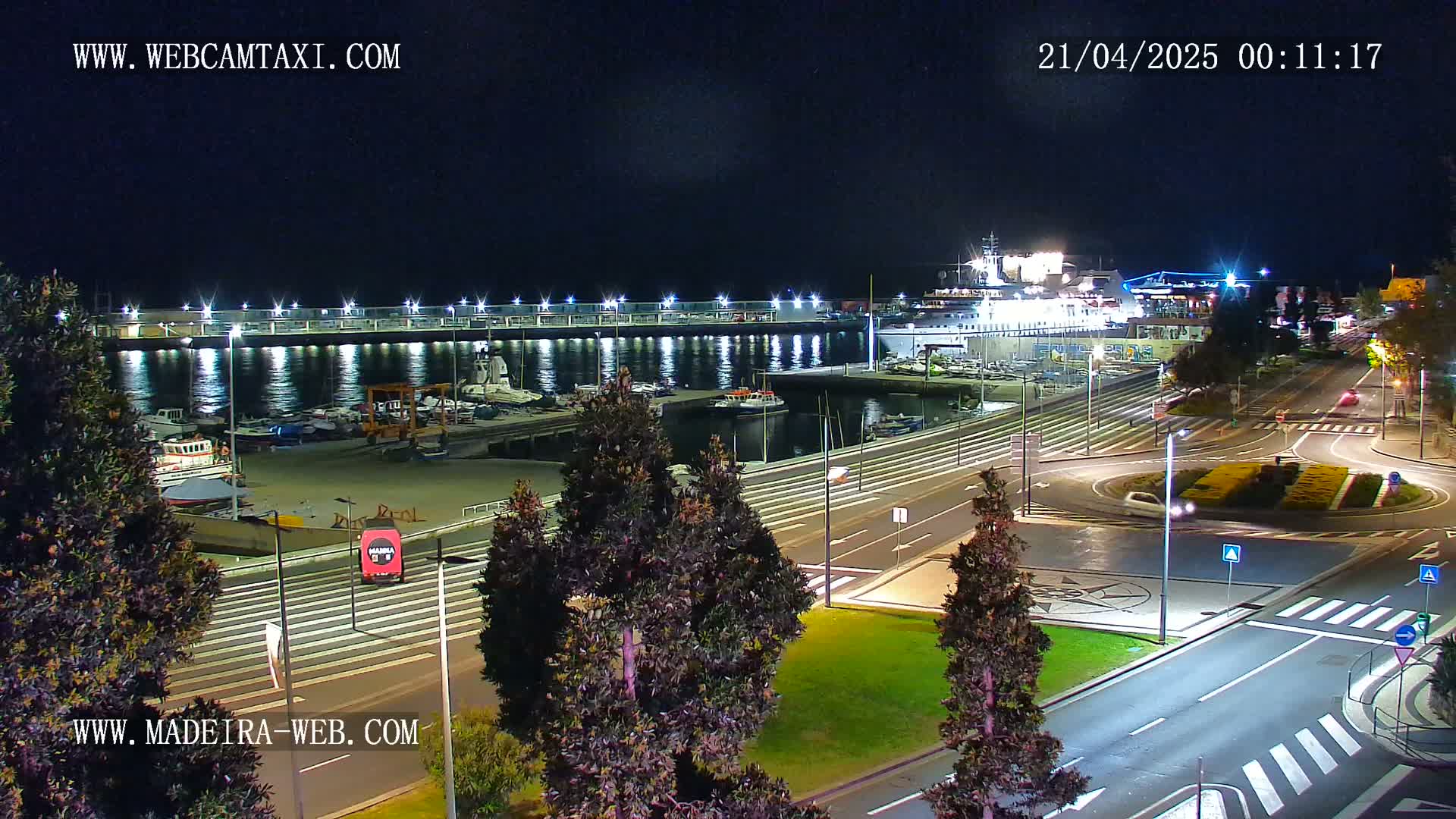 A nighttime view of a harbor with docked boats and a large ship, adjacent to well-lit roads and a roundabout, surrounded by dark green trees.
