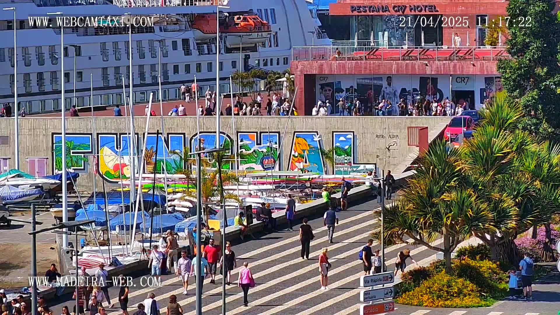 A sunny harbor scene shows a large cruise ship docked near a brightly colored mural of the word "FUNCHAL," with numerous people walking along a promenade and many small boats moored nearby.