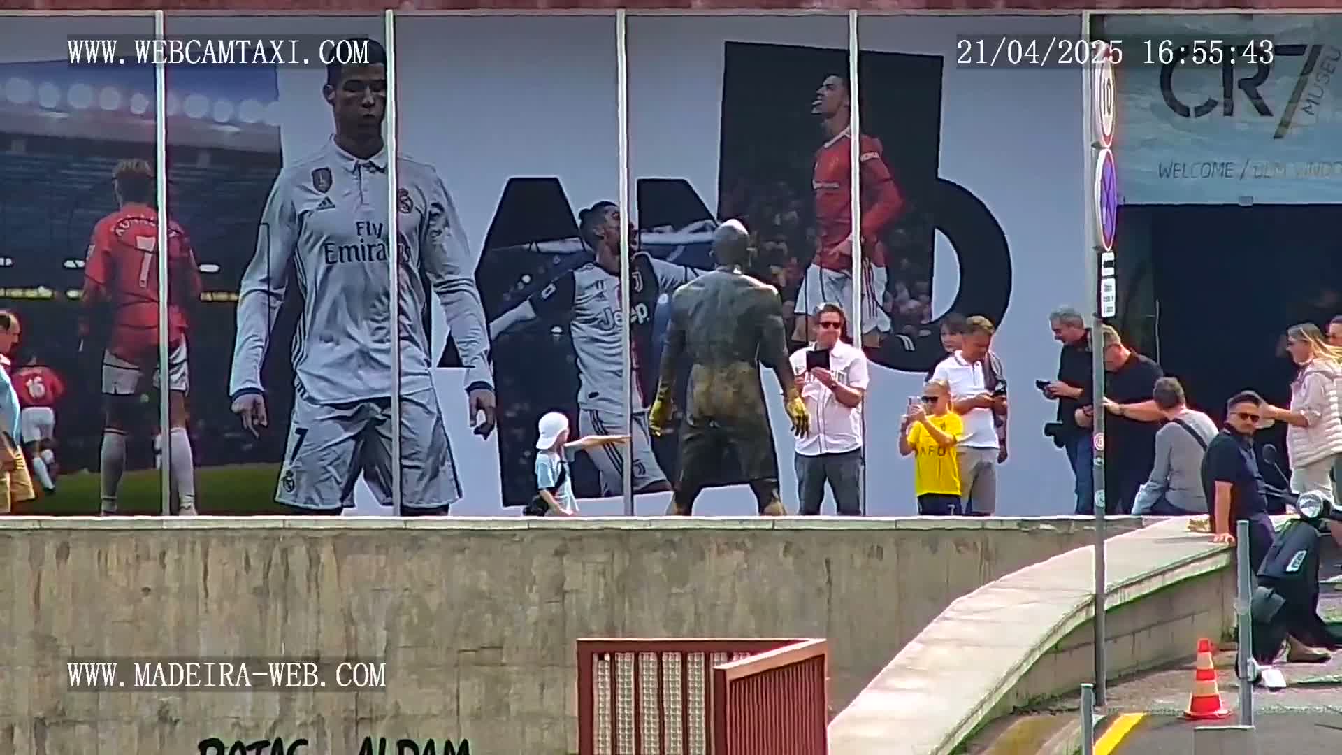 A group of people stand outside on a sunny day in front of a large wall displaying images of Cristiano Ronaldo and other soccer players.