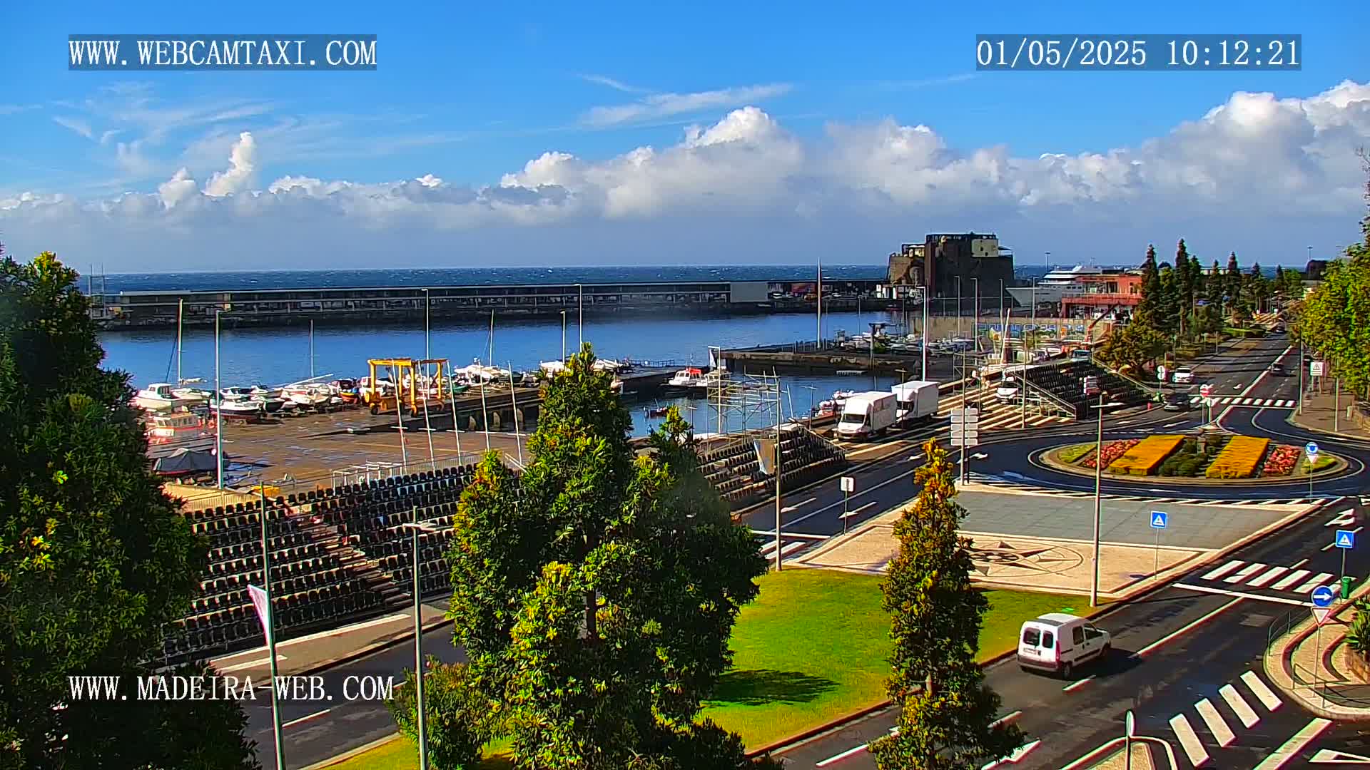 A harbor filled with boats is visible from an elevated vantage point on a partly cloudy day, with a roadway and roundabout in the foreground.