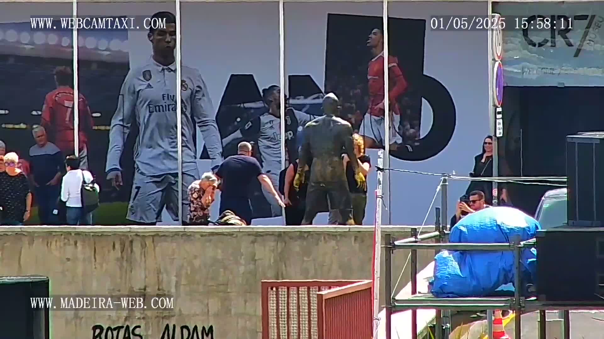 A group of people stand near a large billboard depicting soccer players, while a bronze statue is unveiled on a sunny day.