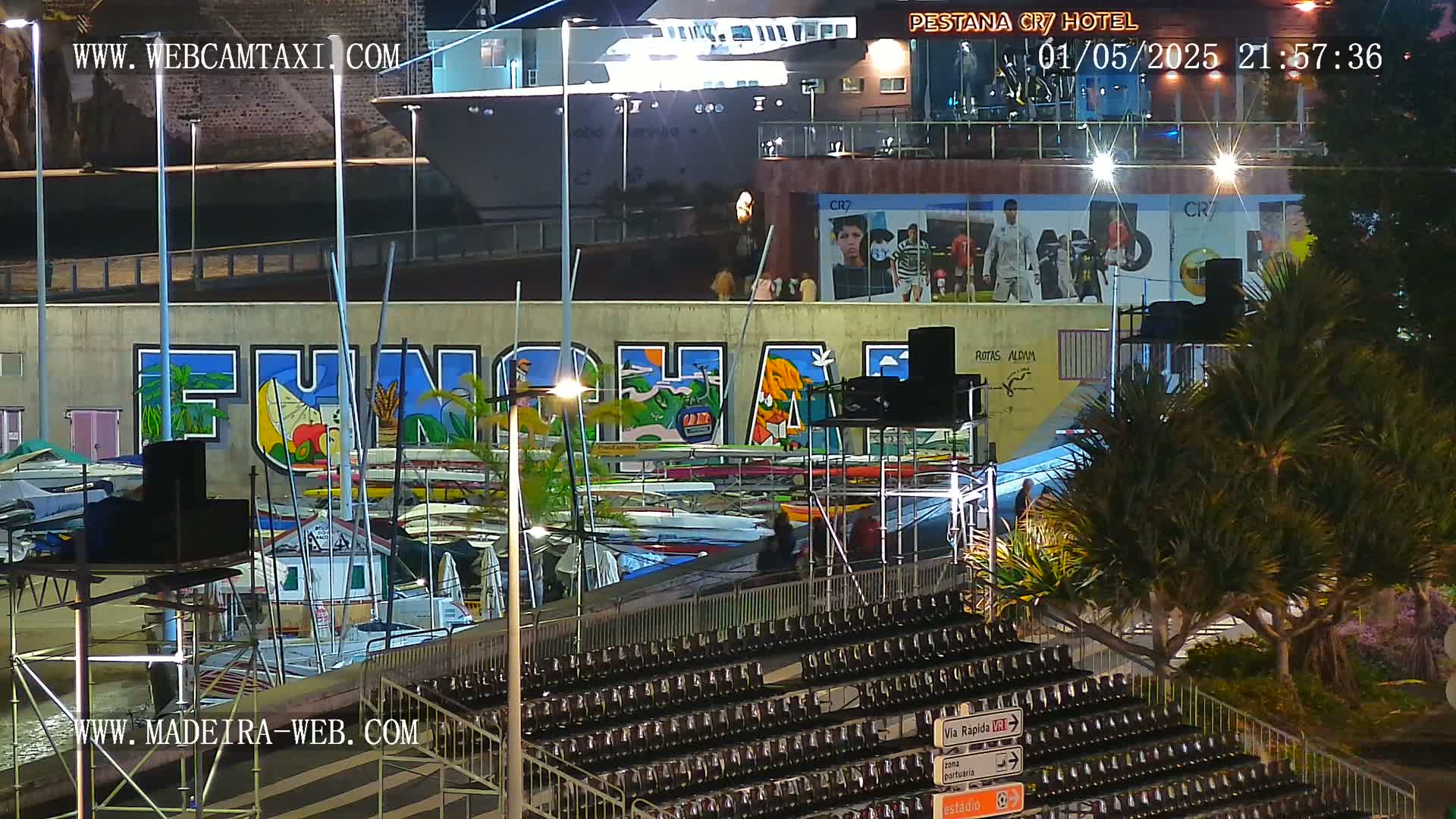 A nighttime view of a harbor with numerous boats docked, a large mural on a wall, empty stadium seating, and palm trees, under artificial light.