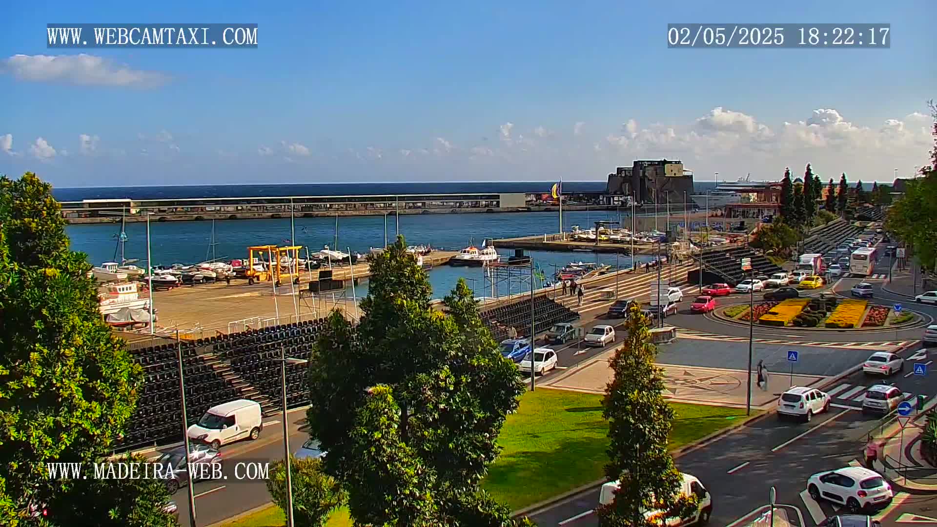 A harbor filled with boats is seen from an elevated vantage point on a sunny day with some clouds, with a roadway and roundabout full of cars nearby.