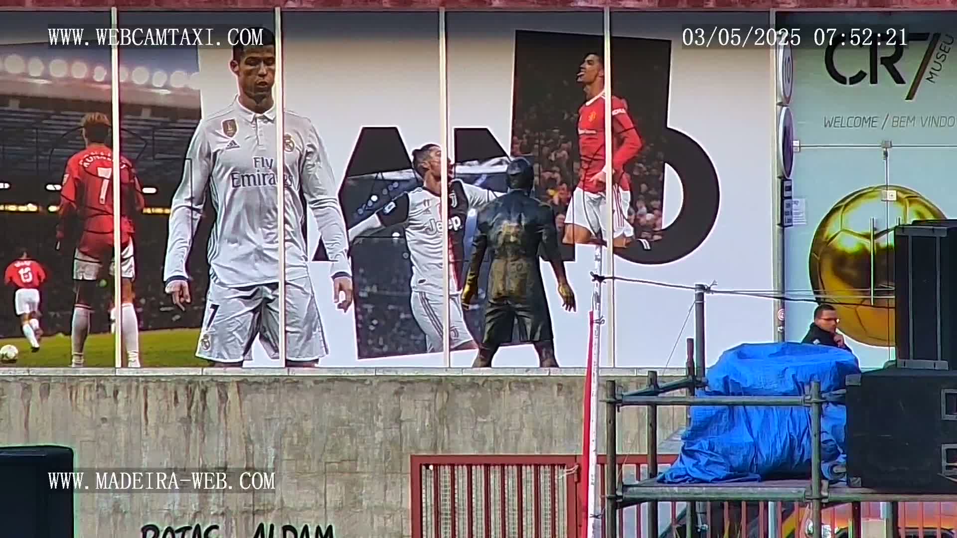A large outdoor billboard displays images of Cristiano Ronaldo in various soccer uniforms, alongside a statue of him, and a person is standing near some equipment on a stage in front of it.