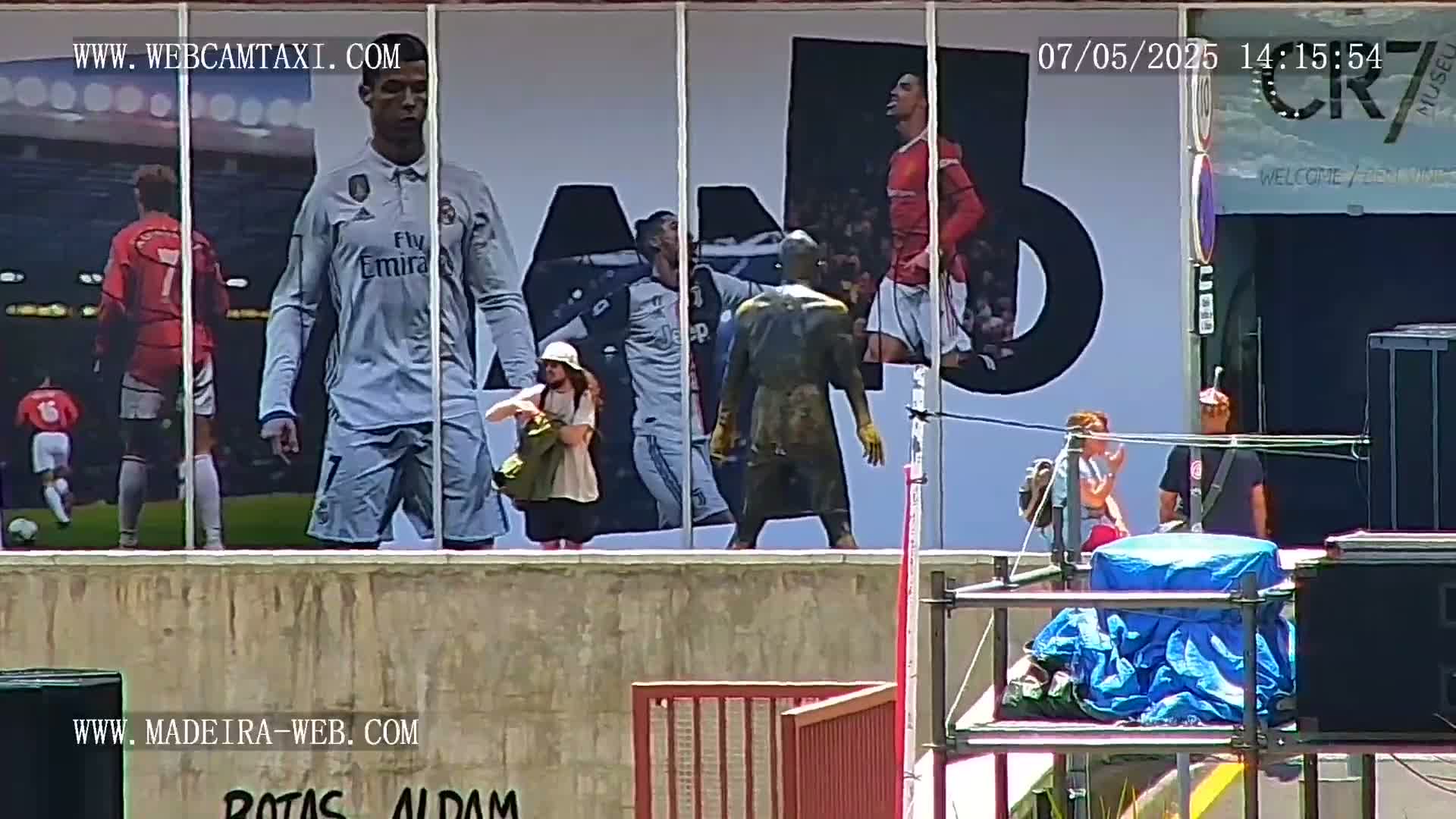 A large mural depicting soccer players is on a building, and several people are standing outside in front of it on a sunny day.