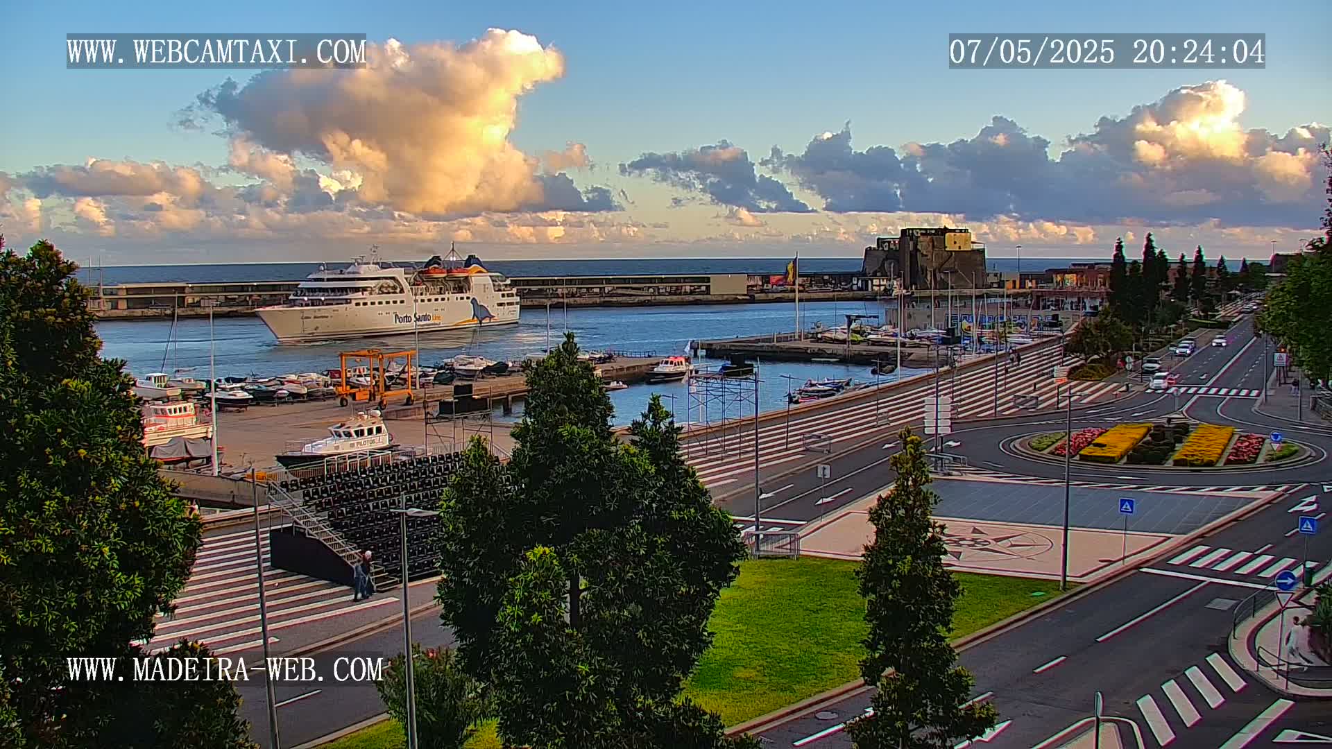 A harbor scene at dusk shows a large ferry docked near many smaller boats, with a paved area, a roundabout with flowers, and a road leading away from the waterfront under a partly cloudy sky.