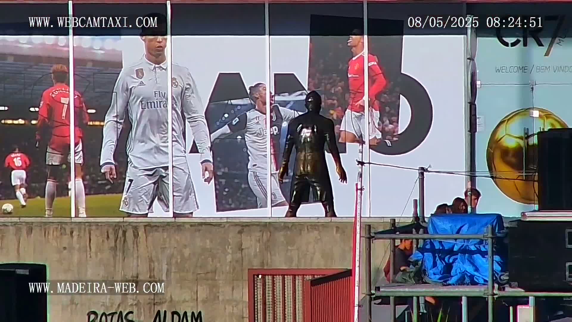 A dark statue of a soccer player stands in front of a large billboard displaying images of Cristiano Ronaldo in various soccer uniforms, under a sunny sky.