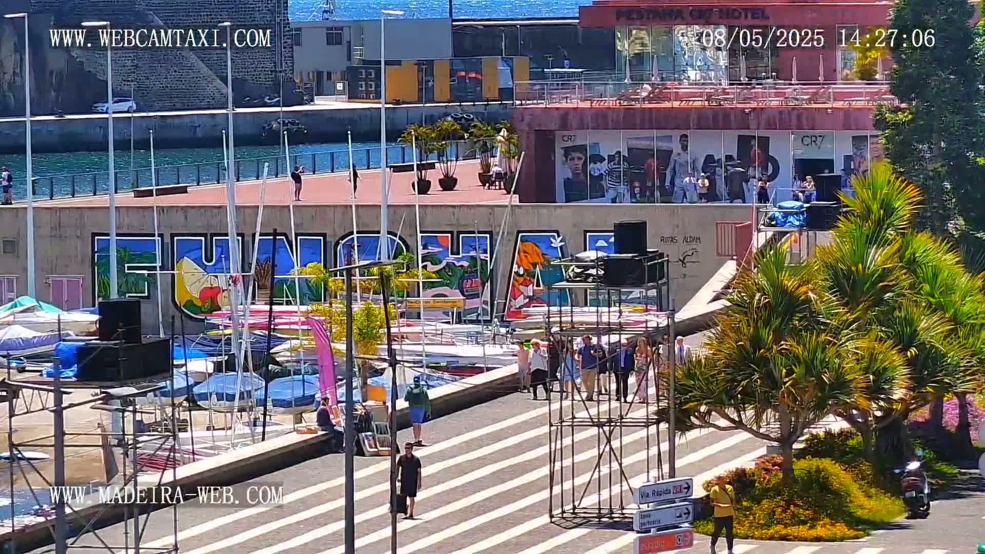 A sunny harbor scene shows numerous sailboats docked, a large mural on a building, and people walking along a paved area with palm trees.