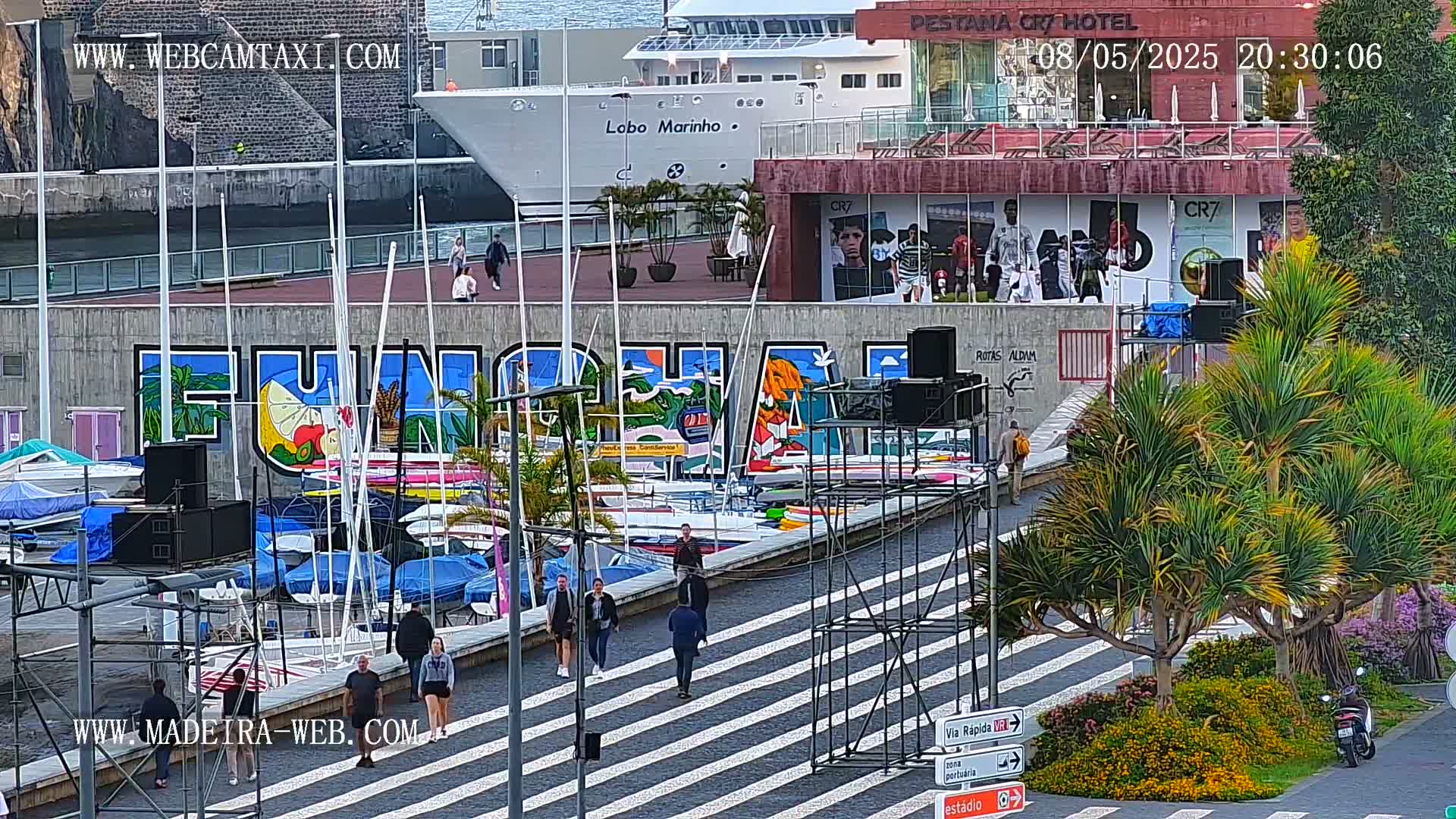 A harbor scene with a large mural of "Funchal," numerous sailboats docked, pedestrians walking along a striped walkway, and lush green palm trees under a partly cloudy sky.