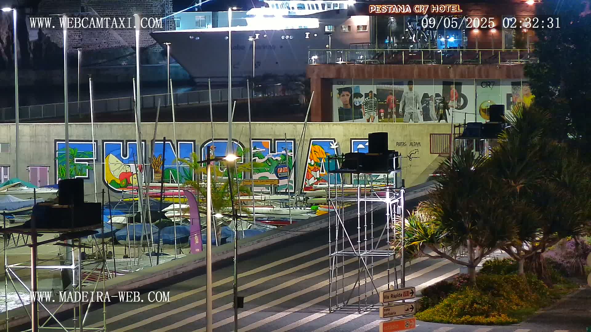 A nighttime harbor scene shows a large mural, numerous boats docked, a large ship partially visible, and some scaffolding near a roadway.
