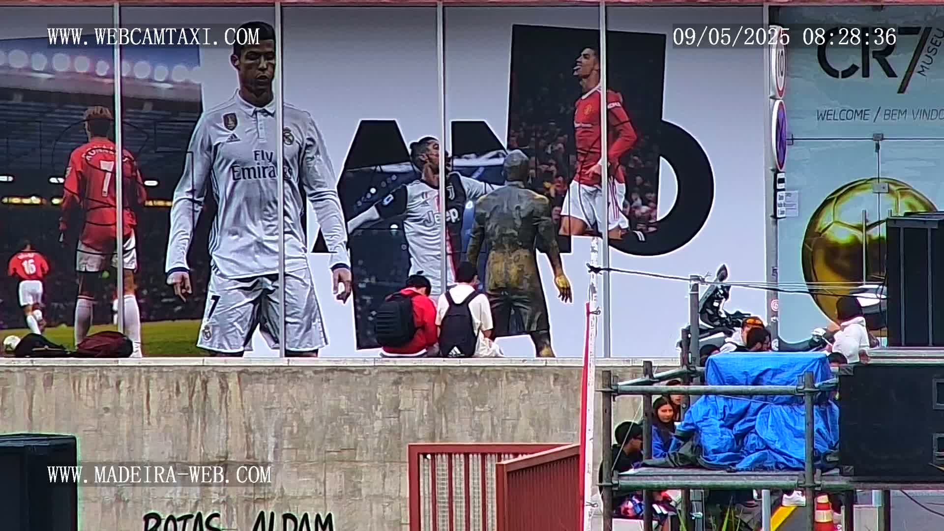 A large outdoor mural depicting Cristiano Ronaldo in various soccer uniforms is partially obscured by several people standing on a platform in front of it.