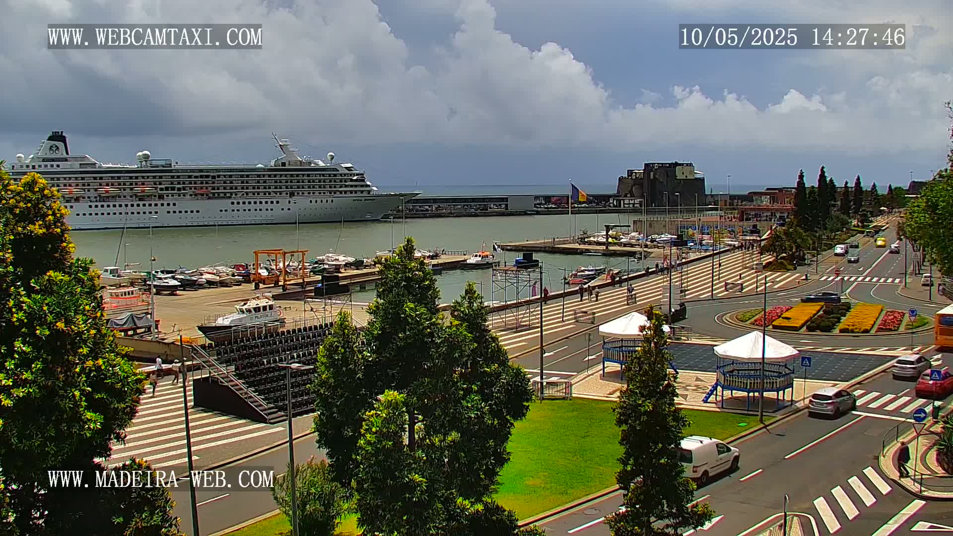 A large cruise ship is docked at a harbor, with a paved walkway and several cars visible along a waterfront road under partly cloudy skies.