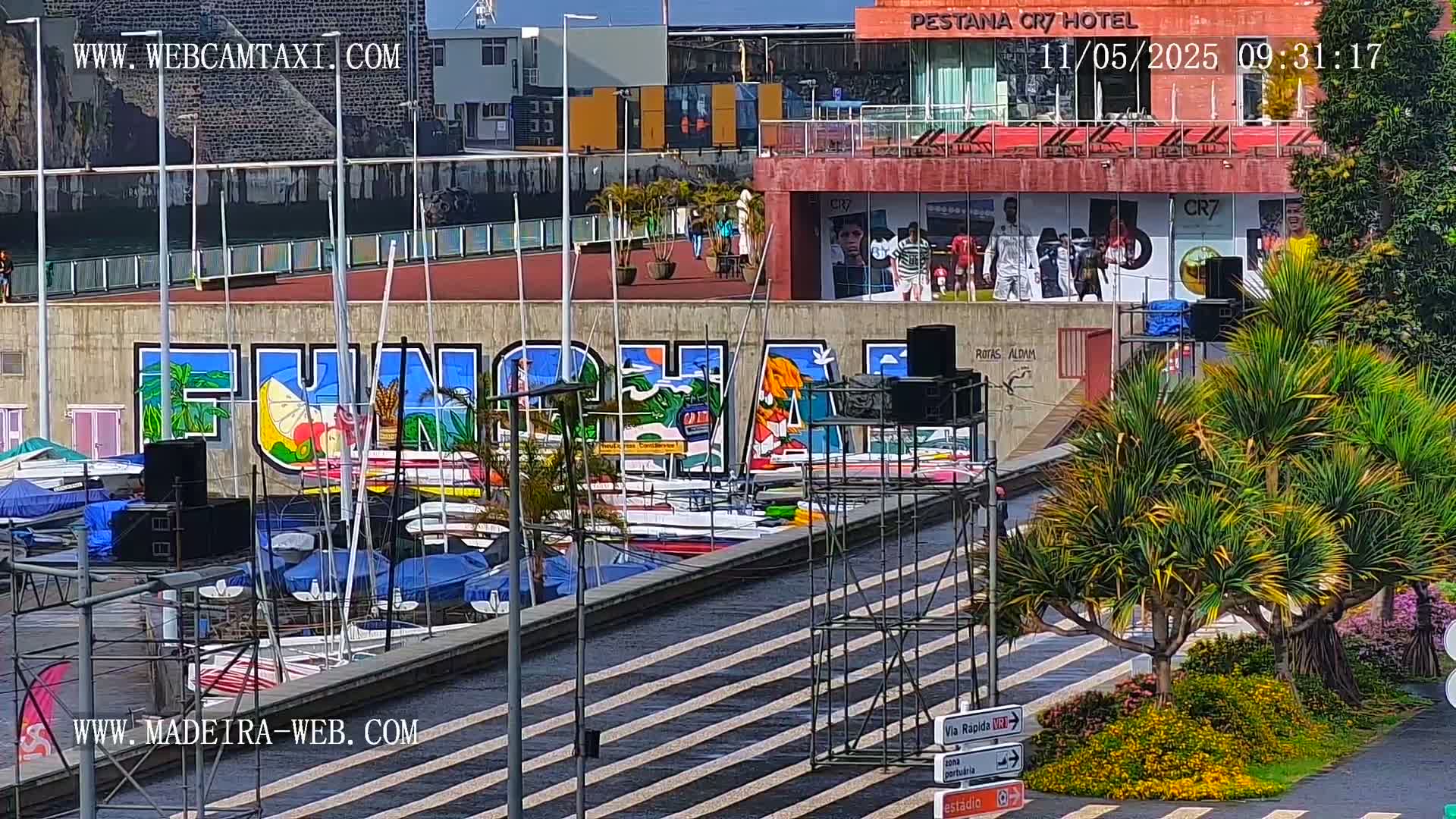 A marina filled with sailboats is situated below a colorful mural and a hotel on a sunny day.