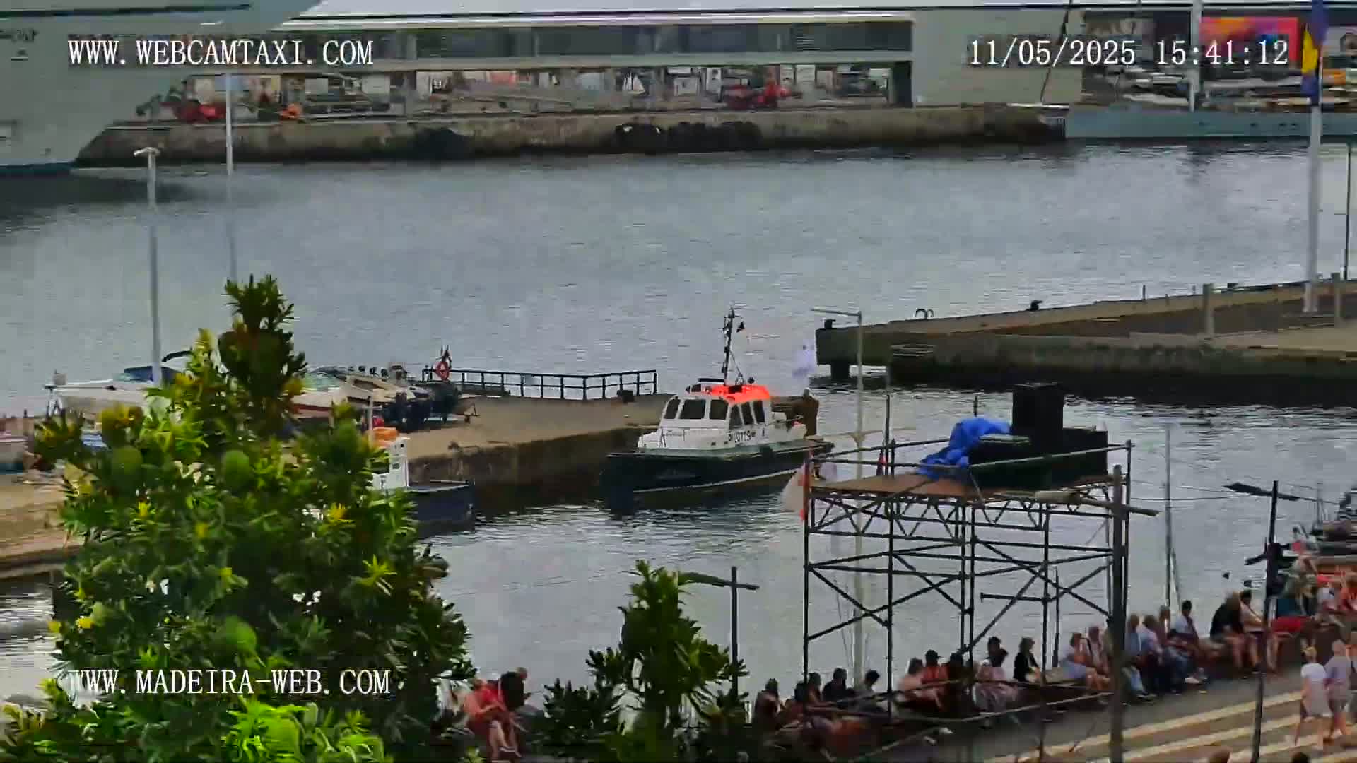 A harbor scene shows a small boat near a dock with several people sitting on the shoreline under a partly cloudy sky.