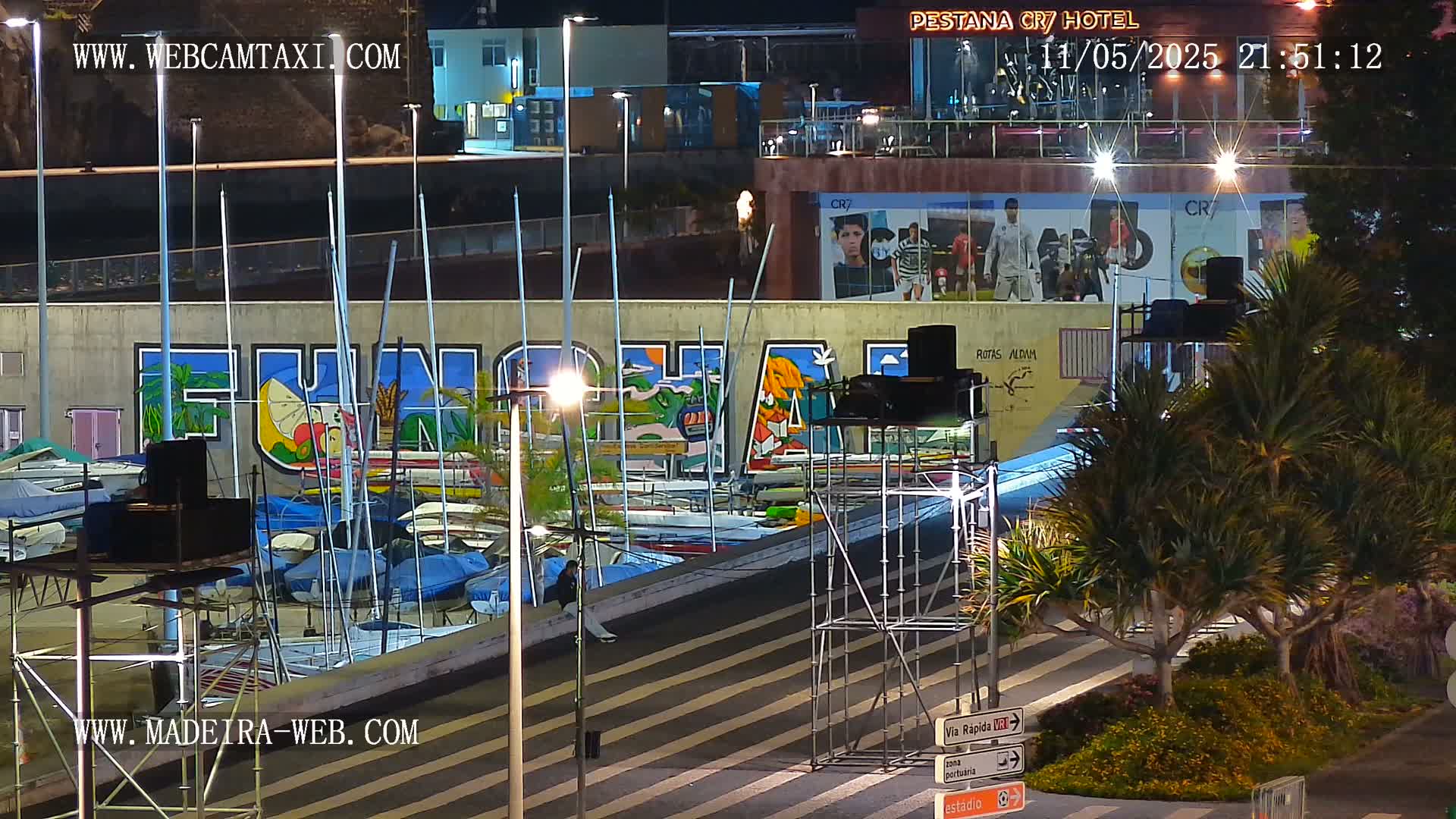 A nighttime view of a harbor with numerous sailboats docked, a large mural depicting the word "Funchal" and tropical imagery on a wall behind it, and palm trees lining a street in front.