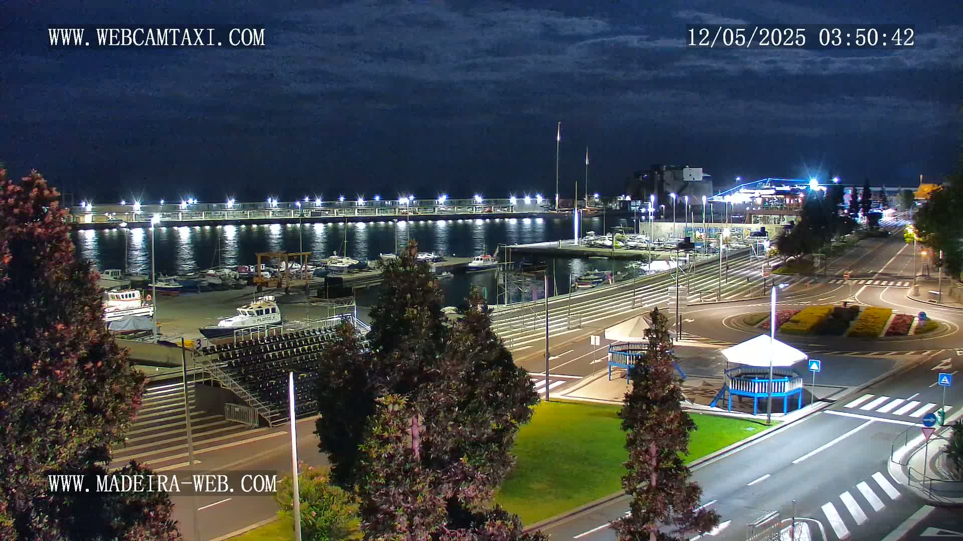 A nighttime view of a marina filled with boats, adjacent to a plaza with roadways and landscaping under a partly cloudy sky.