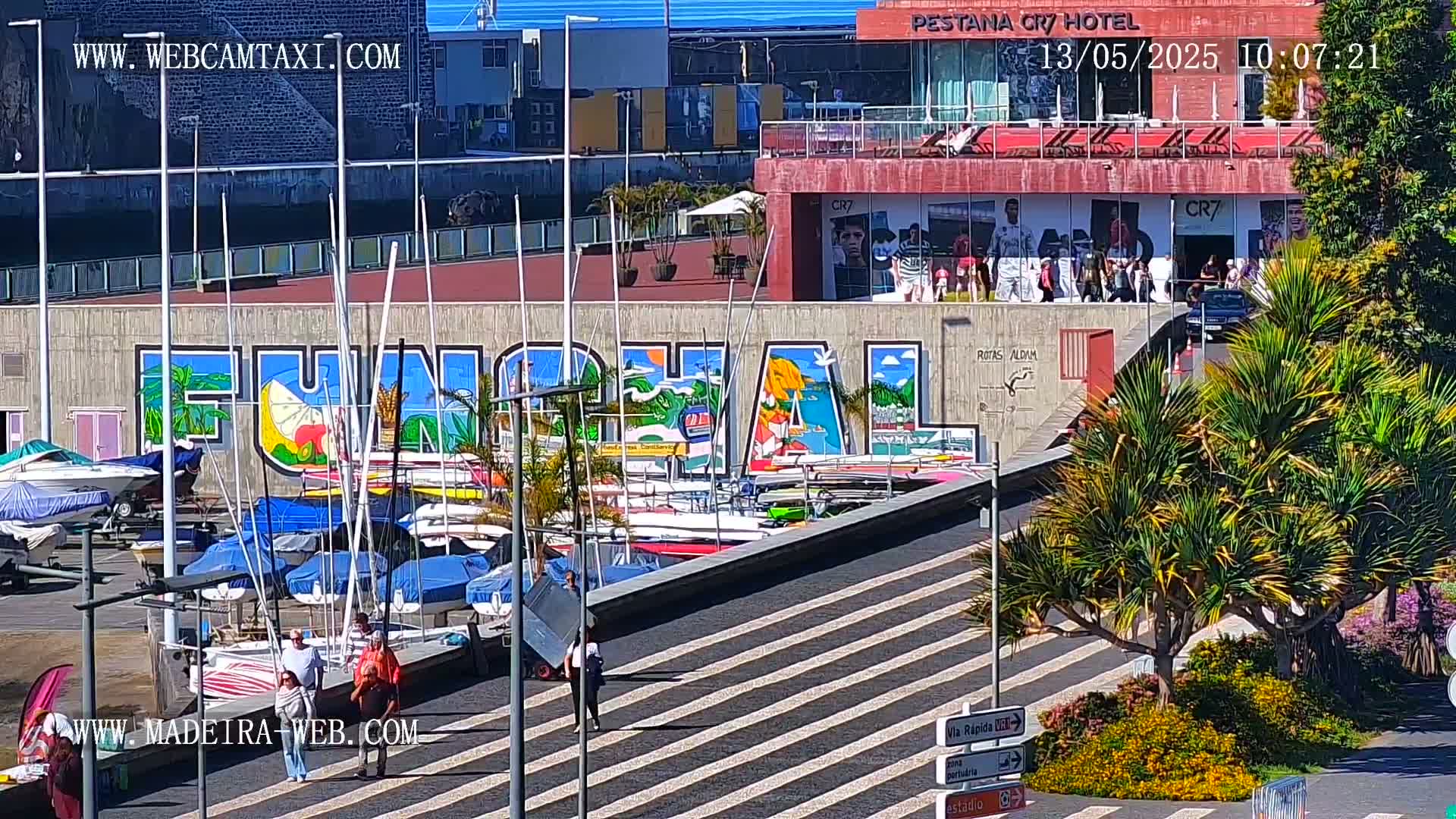 A sunny harbor scene shows numerous sailboats docked next to a large mural of the word "FUNCHAL," with a hotel and people visible in the background.