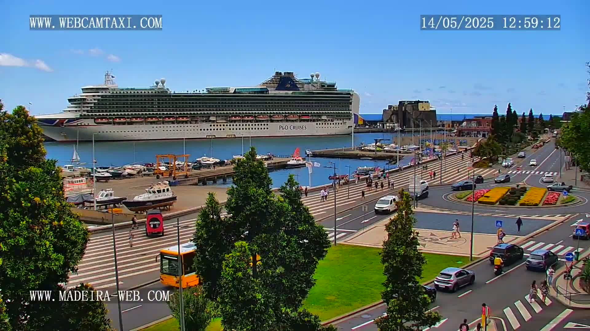 A large cruise ship is docked at a harbor on a sunny day, with pedestrians and cars visible on a nearby roadway and in a roundabout.