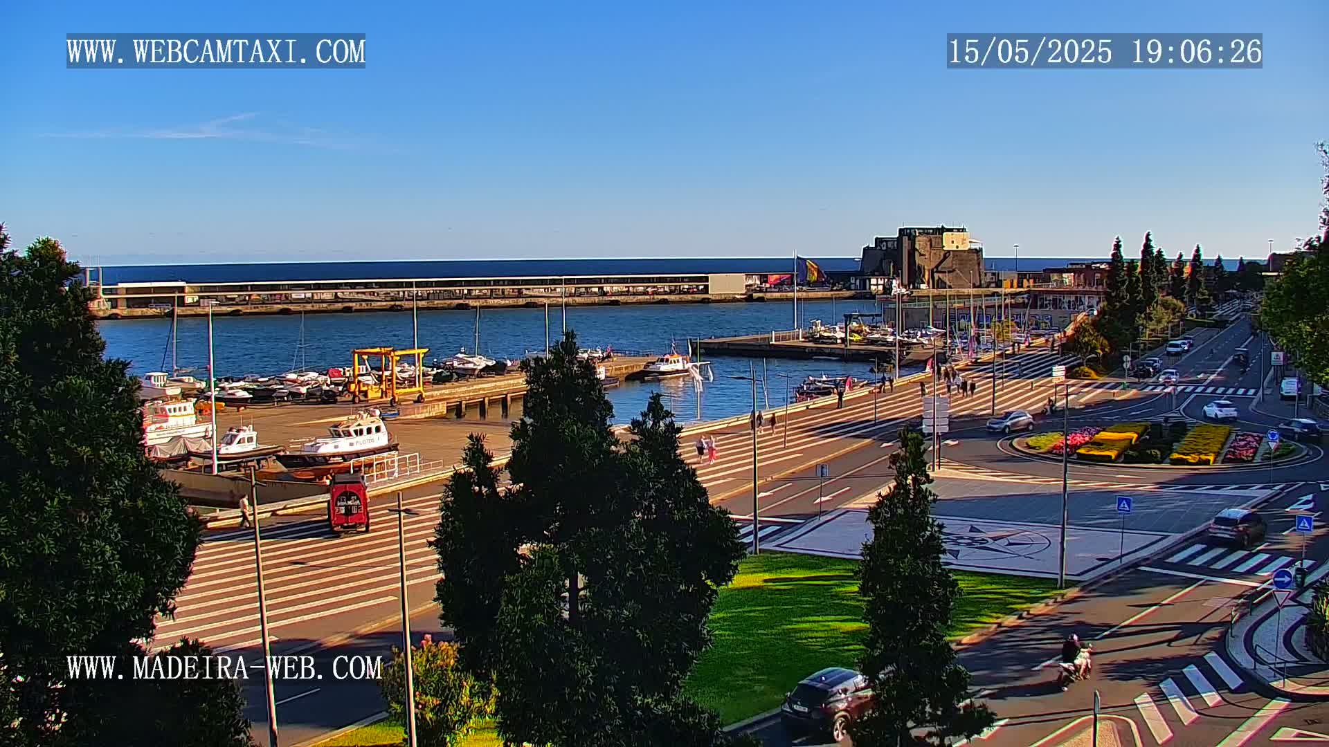 A harbor filled with boats is seen from an elevated vantage point on a sunny day, with a paved area and roadways leading to a roundabout decorated with flowers.