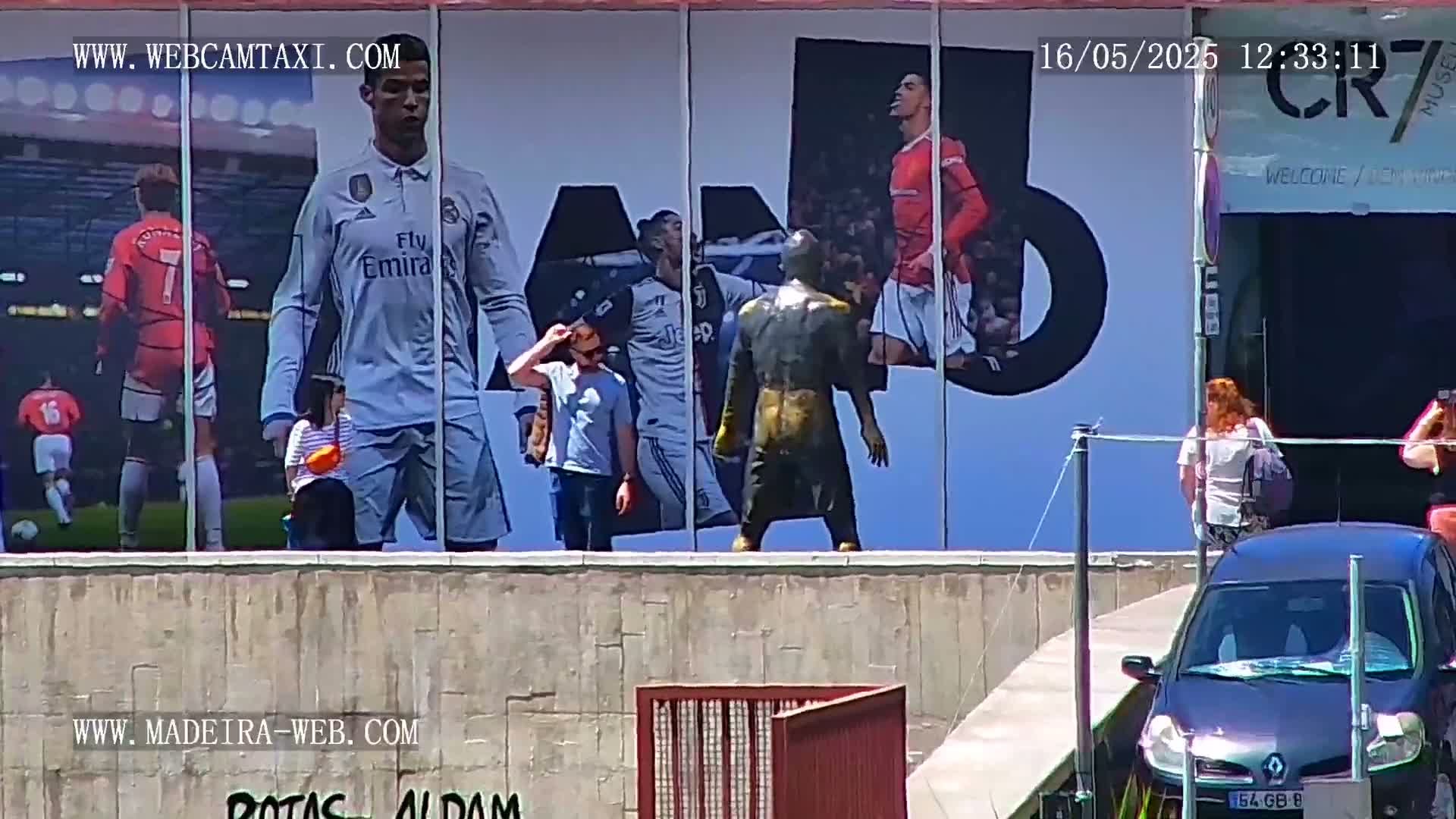 A large billboard displaying images of Cristiano Ronaldo in different soccer uniforms is visible in bright, sunny weather, with several people standing in front of it and a car parked nearby.
