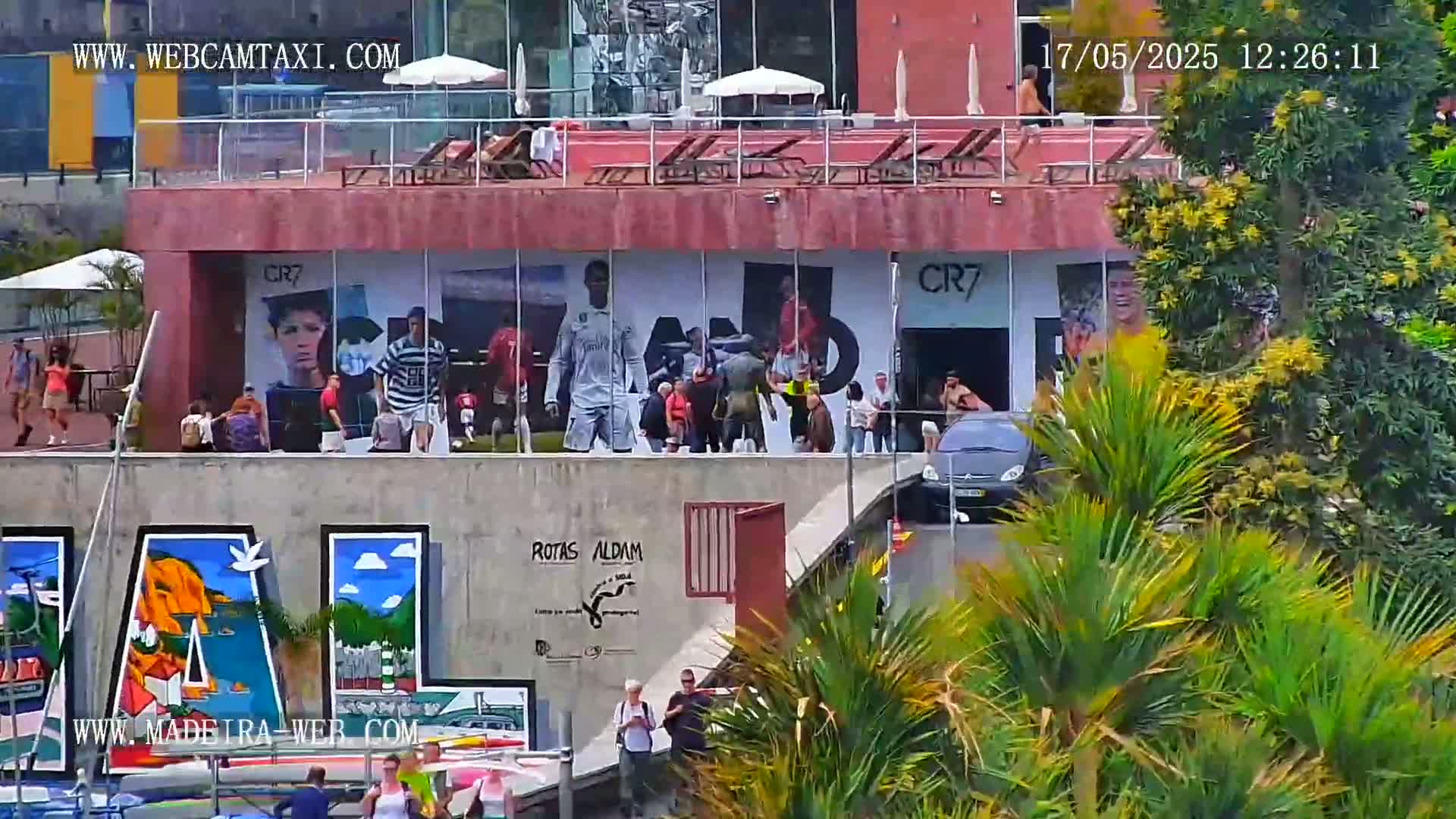 A building with large images of soccer players is surrounded by people and lush greenery on a sunny day.