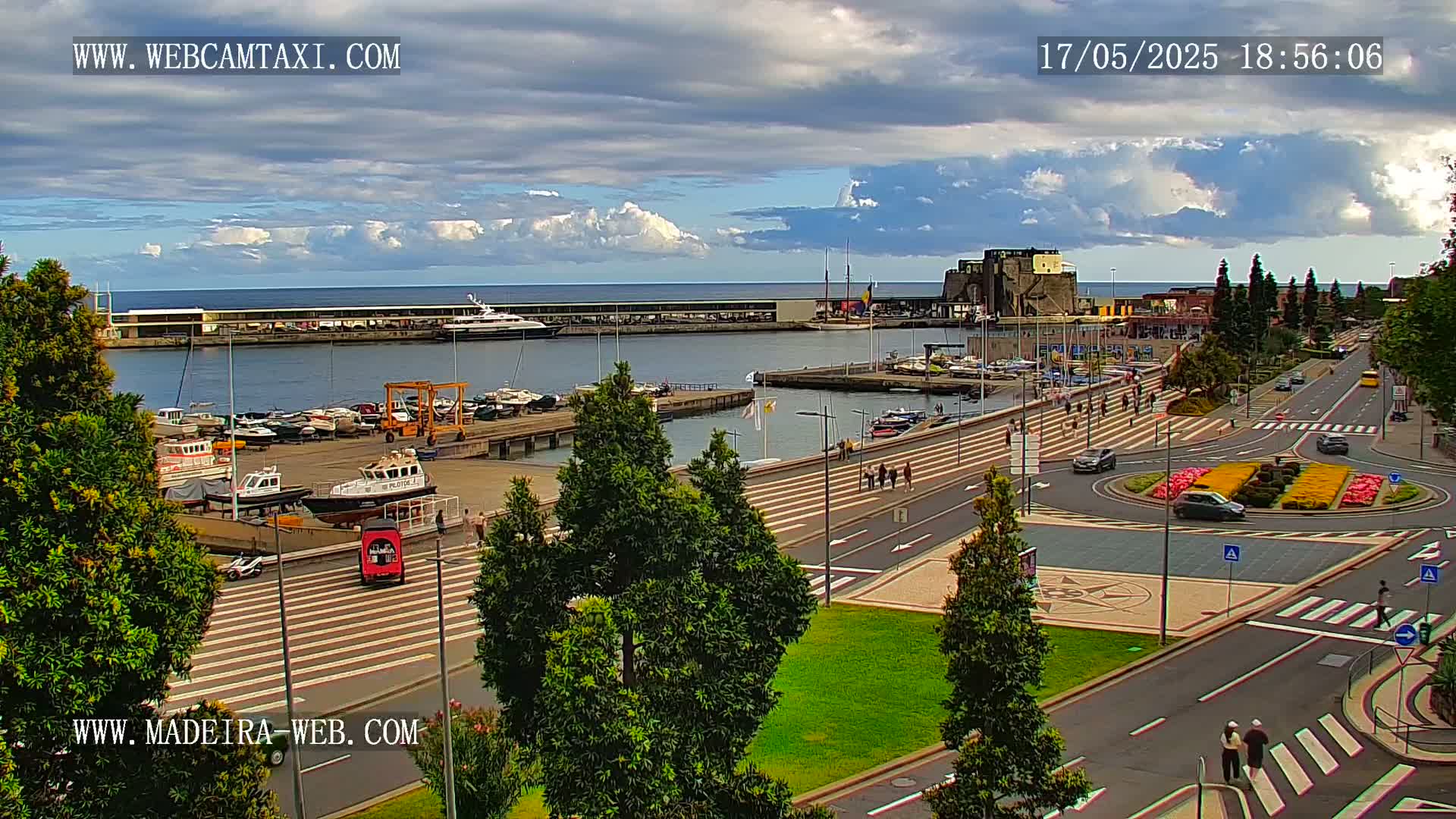 A harbor filled with boats and yachts is visible under a partly cloudy sky, with a paved area and roads leading to a roundabout with landscaping in the foreground.