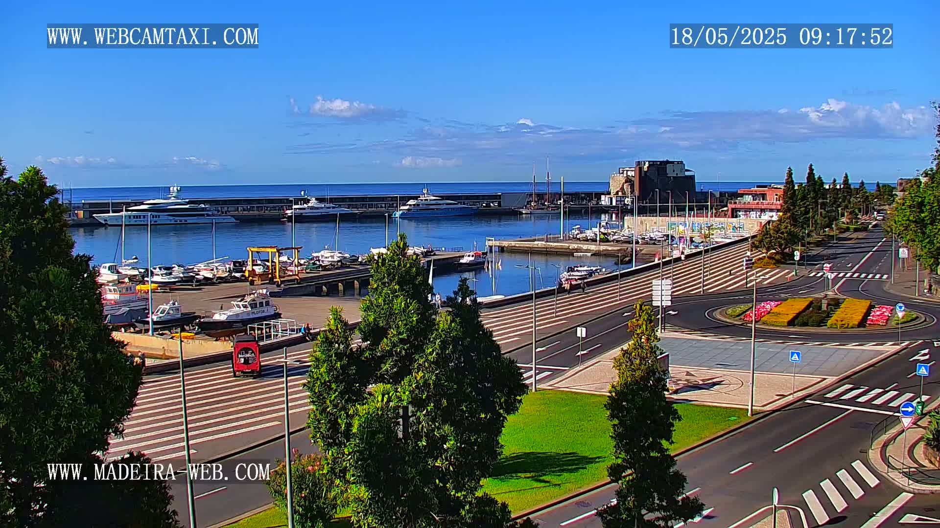 A marina filled with boats sits on a calm ocean under a mostly sunny sky, next to a paved area with green landscaping and roadways.