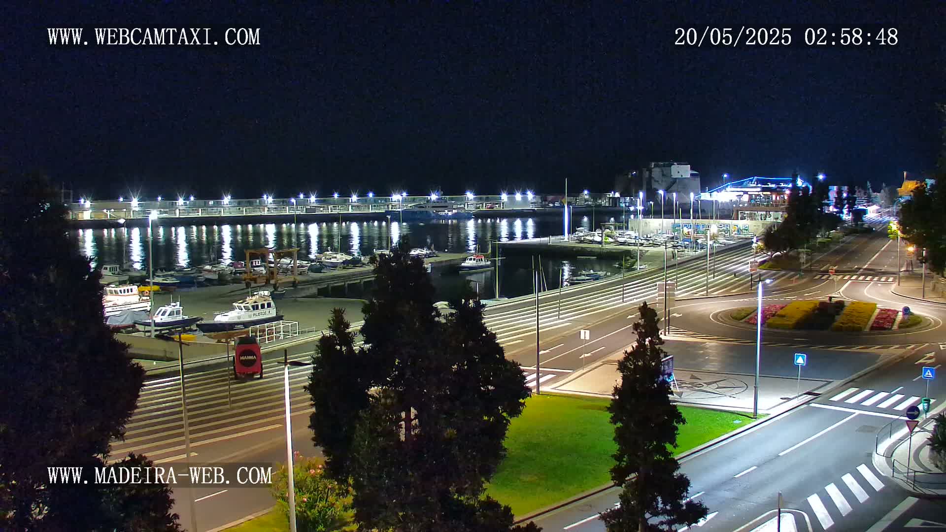 A nighttime view of a harbor with boats docked, illuminated by lights, next to a roadway and roundabout.