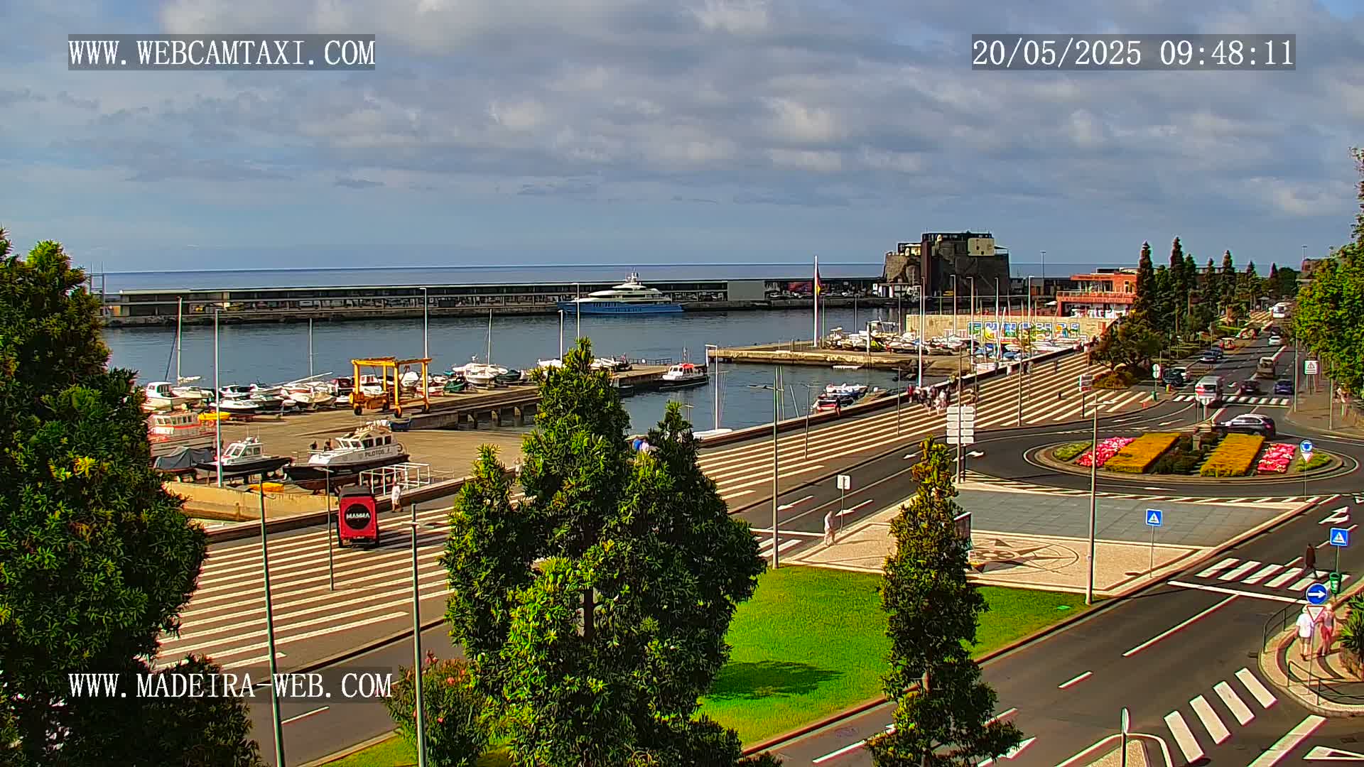 A harbor filled with boats is visible from an elevated vantage point overlooking a paved plaza with a roundabout and several cars under partly cloudy skies.