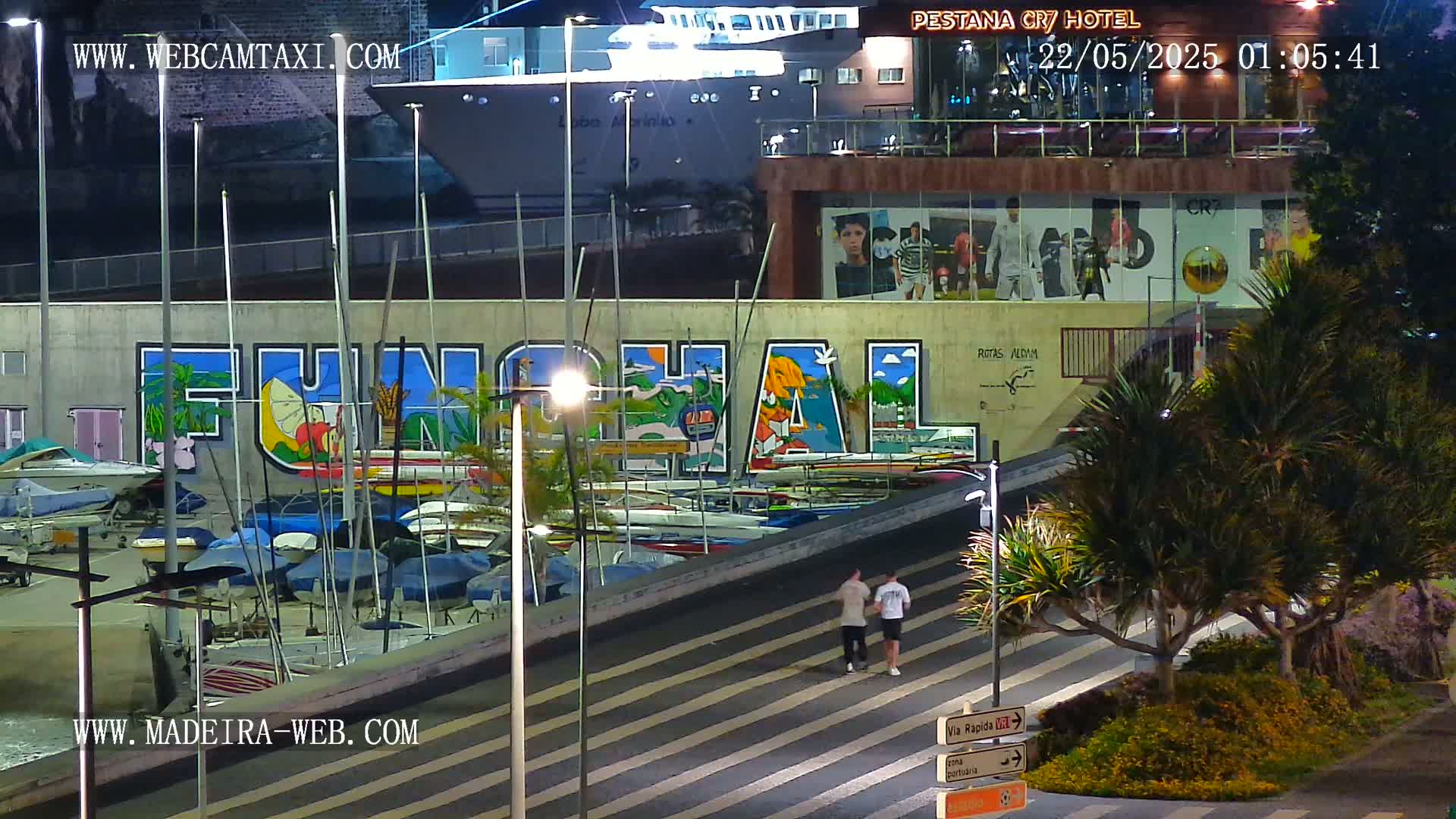 At night, a harbor scene shows a large mural of the word "Funchal" on a building next to a marina filled with boats, with two people walking along a nearby roadway under illuminated streetlights.