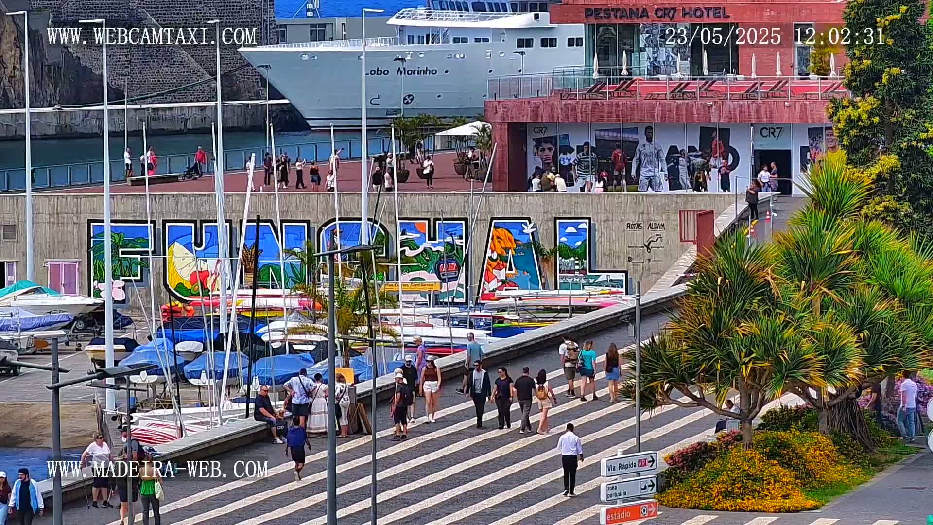 A sunny harbor scene shows a large mural of the word "Funchal" on a wall next to a marina filled with sailboats, with pedestrians walking on a crosswalk in front of a hotel.