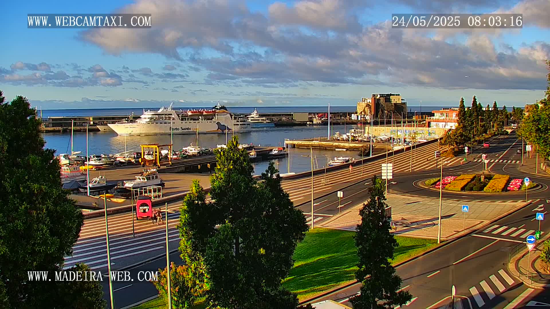 A harbor with a large ferry and numerous smaller boats, situated next to a paved area with a roundabout and green landscaping, under a partly cloudy sky.