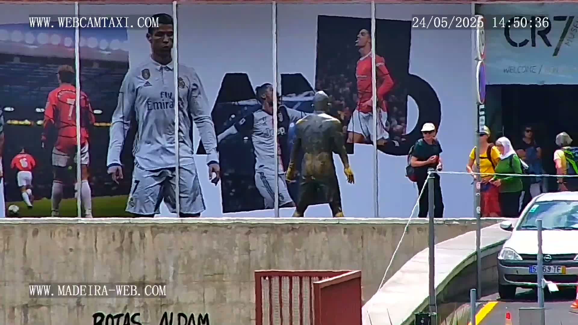 A large building displays images of Cristiano Ronaldo in various soccer uniforms, while several people walk past it on a sunny day.