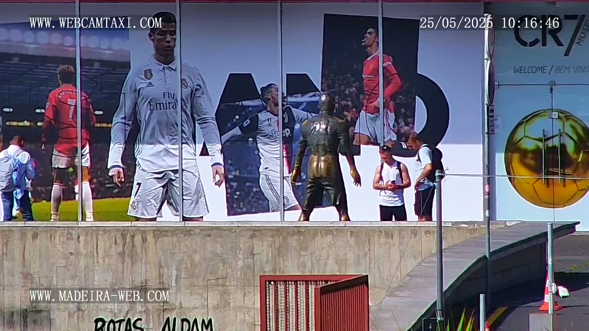 A large outdoor billboard displaying images of Cristiano Ronaldo in different soccer uniforms is viewed from a low angle on a sunny day, with several people standing in front of it.