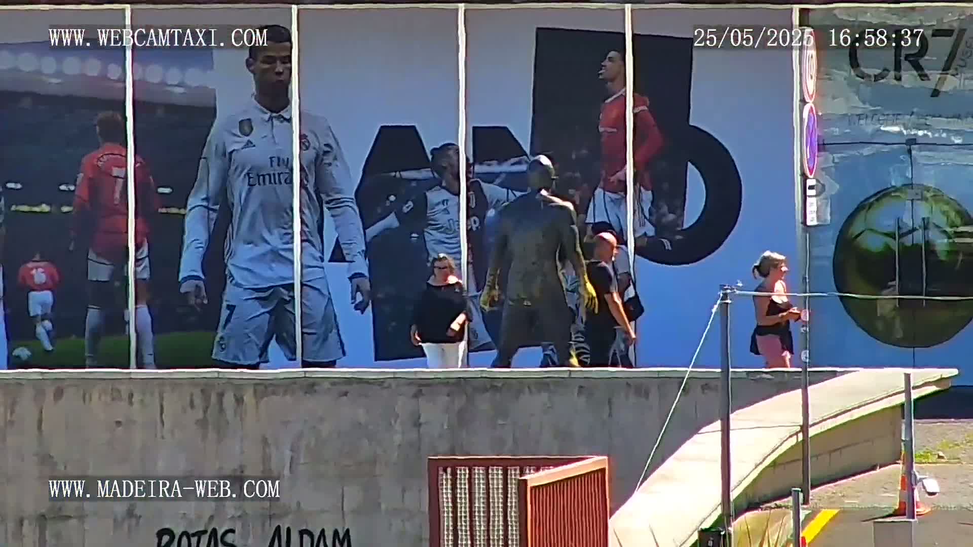 A large billboard depicting soccer players is visible in the sunny outdoor scene, with several people walking past it.