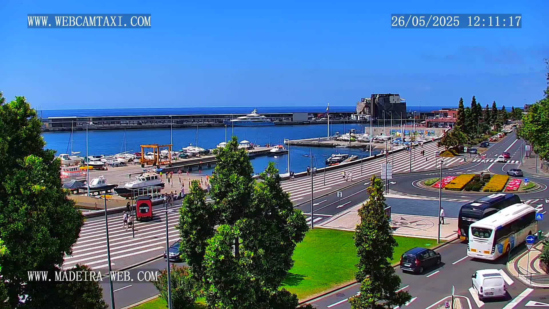 A sunny harbor scene shows a marina filled with boats, a paved waterfront area with pedestrians and vehicles, and a large yacht in the distance.