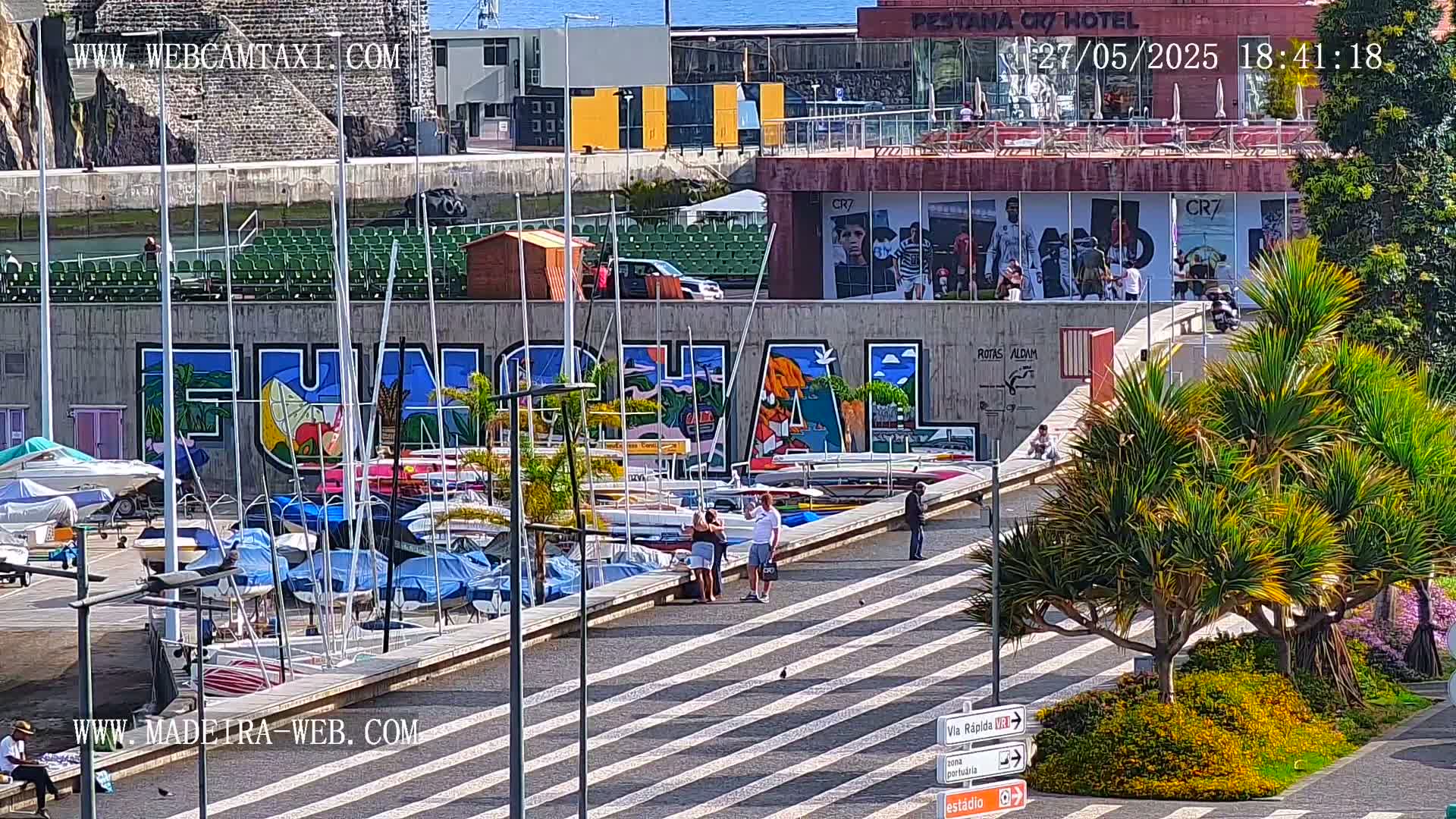 A sunny harbor scene features numerous sailboats docked along a wall with a large mural, several people walking along a paved area, and lush green plants.