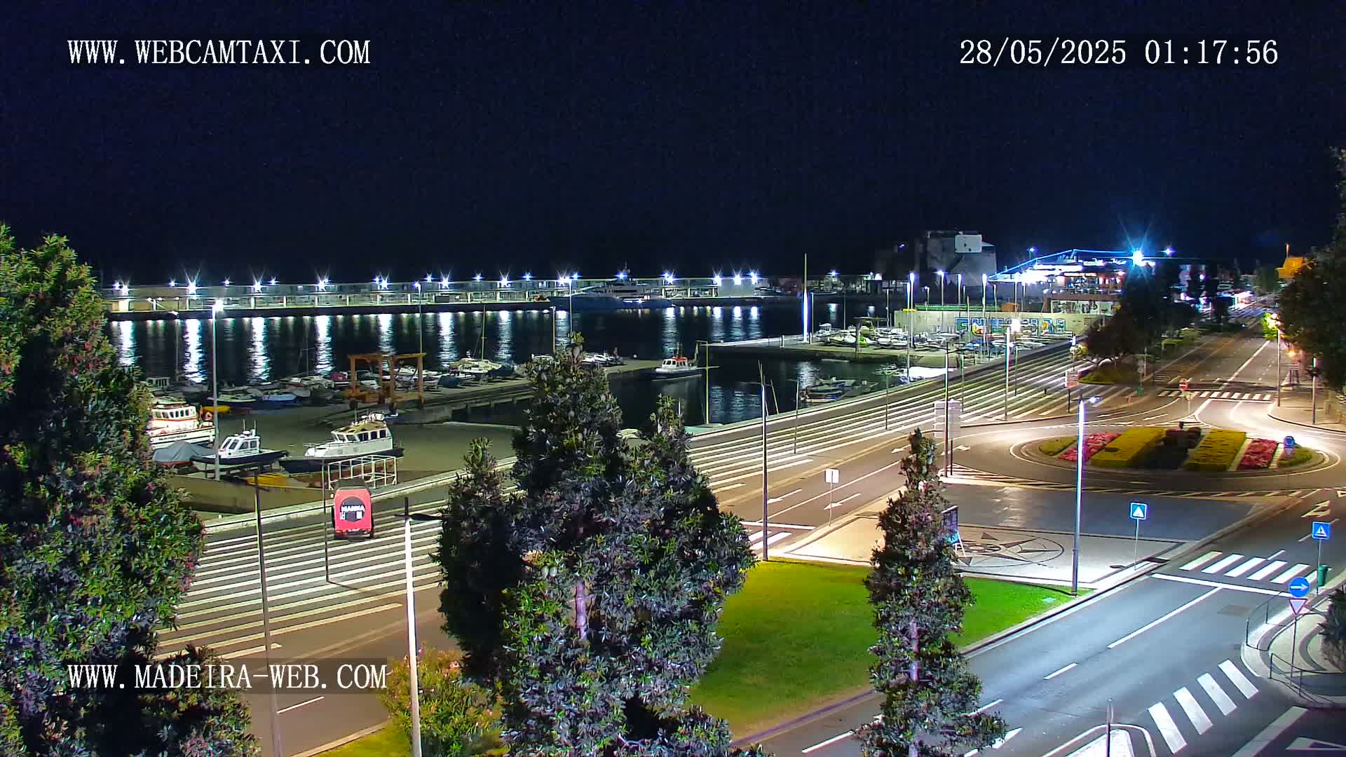 A nighttime view of a harbor filled with boats, adjacent to a roadway with a roundabout, all brightly lit.