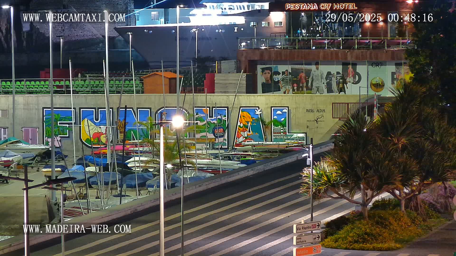 A nighttime harbor scene shows numerous boats docked along a wall with a large mural, illuminated by streetlights, with a large ship visible in the background.