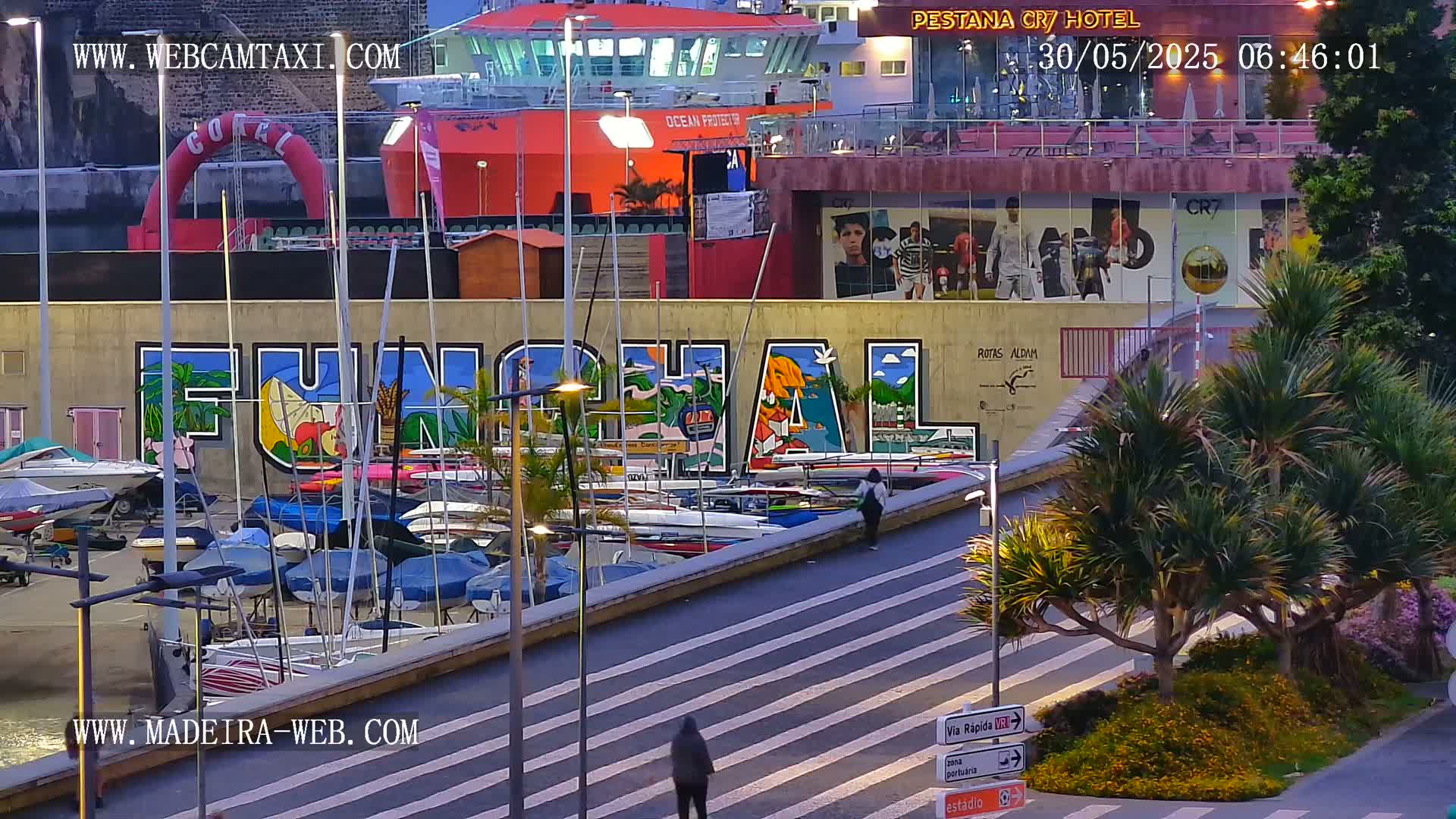 A harbor filled with sailboats is situated below a large mural of the word "FUNCHAL," with a large orange ferry visible in the background under an evening sky.