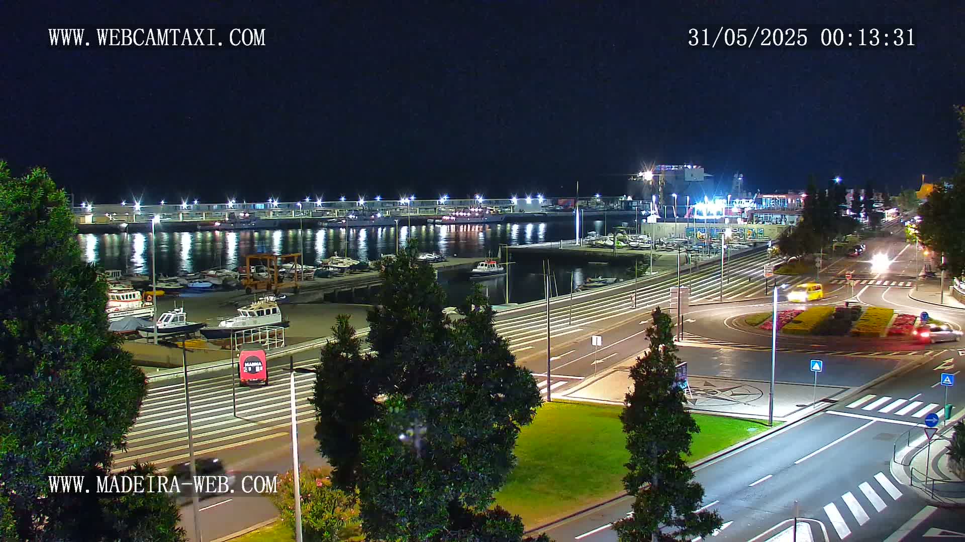 A nighttime aerial view of a harbor filled with boats, adjacent to a roadway with cars and a roundabout, under clear skies.