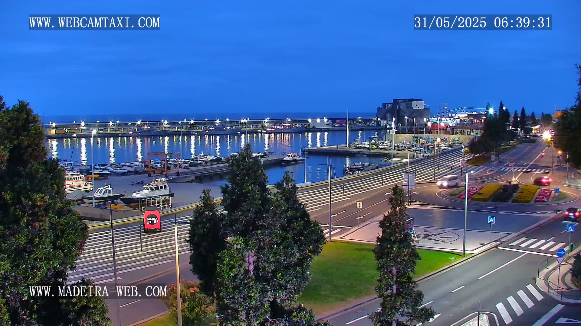 A harbor filled with boats at twilight is visible from an elevated vantage point overlooking a roadway with minimal traffic.