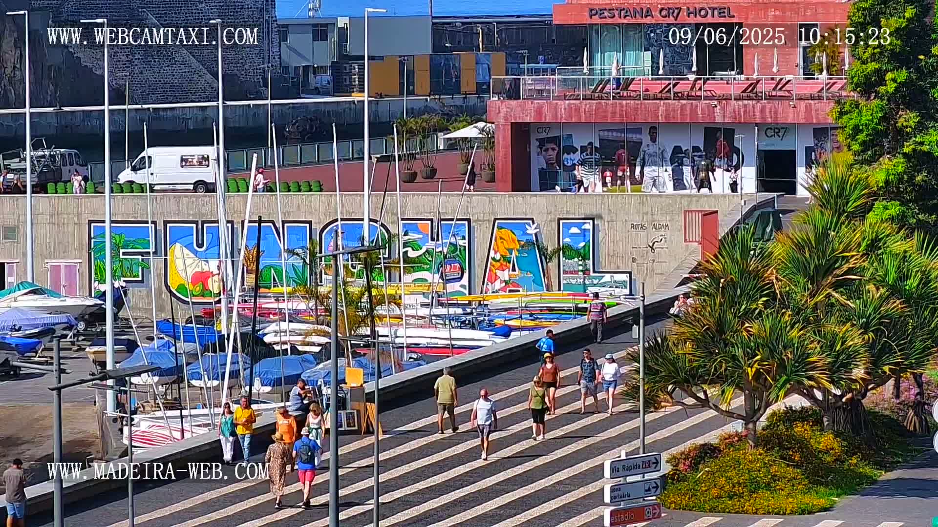 A sunny harbor scene features a large mural of the word "FUNCHAL," numerous small boats docked along a wall, and pedestrians strolling on a crosswalk.