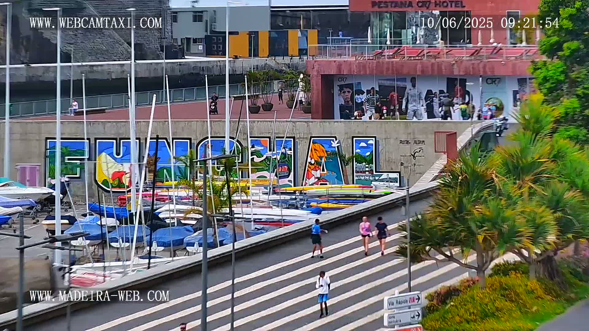 A marina with numerous sailboats and kayaks is situated below a large mural of the word "Funchal" on a wall; several people walk along a nearby pedestrian walkway on a sunny day.