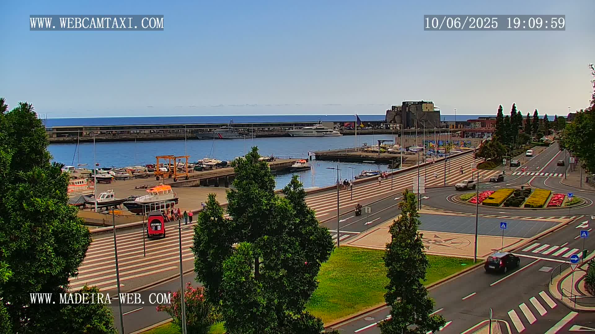 A harbor filled with boats is visible from a waterfront promenade on a sunny day, with cars driving on the roads nearby.