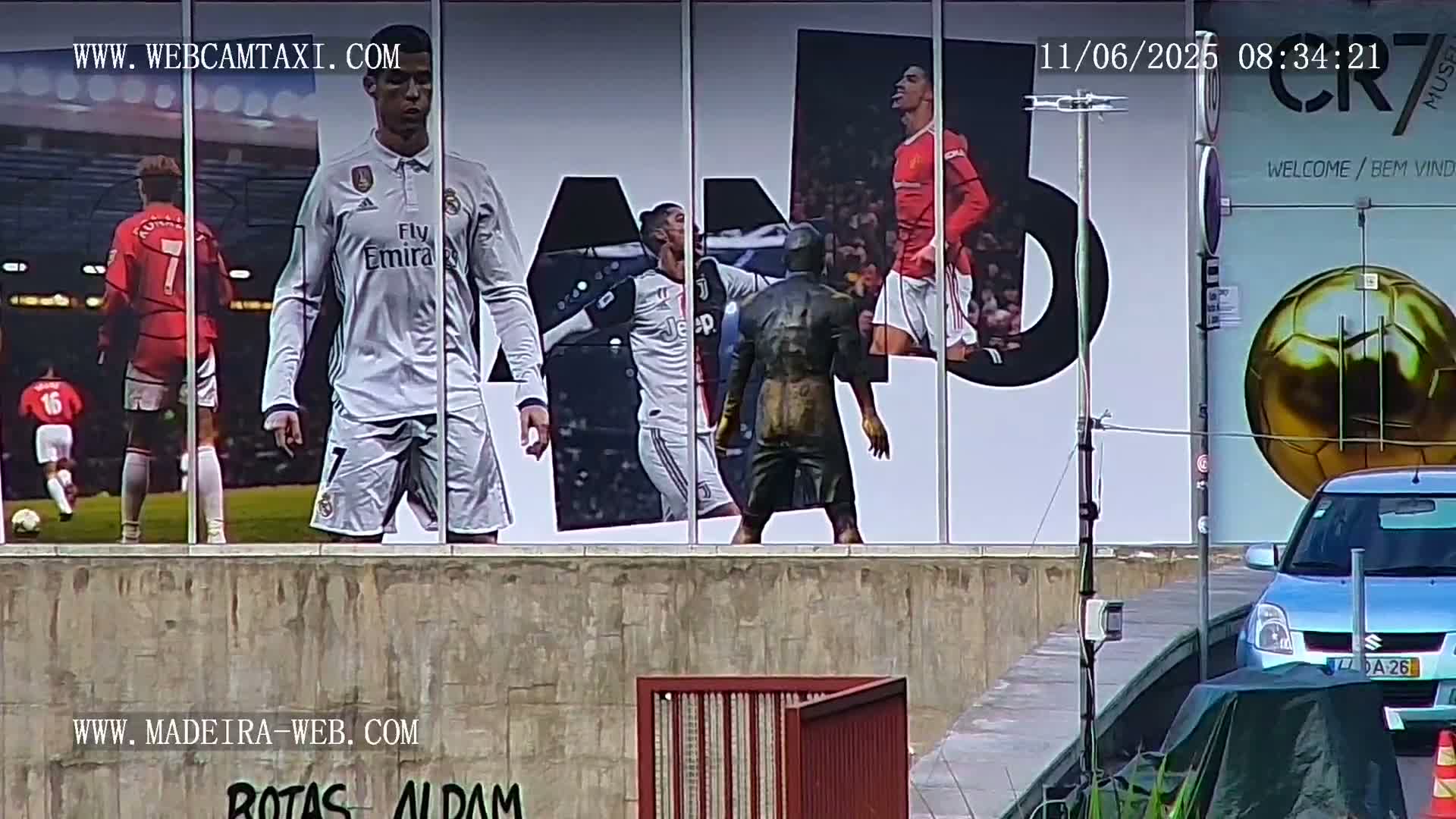 A large billboard featuring images of Cristiano Ronaldo in various soccer uniforms is visible, with a light-blue car parked in front of it on a mostly overcast day.