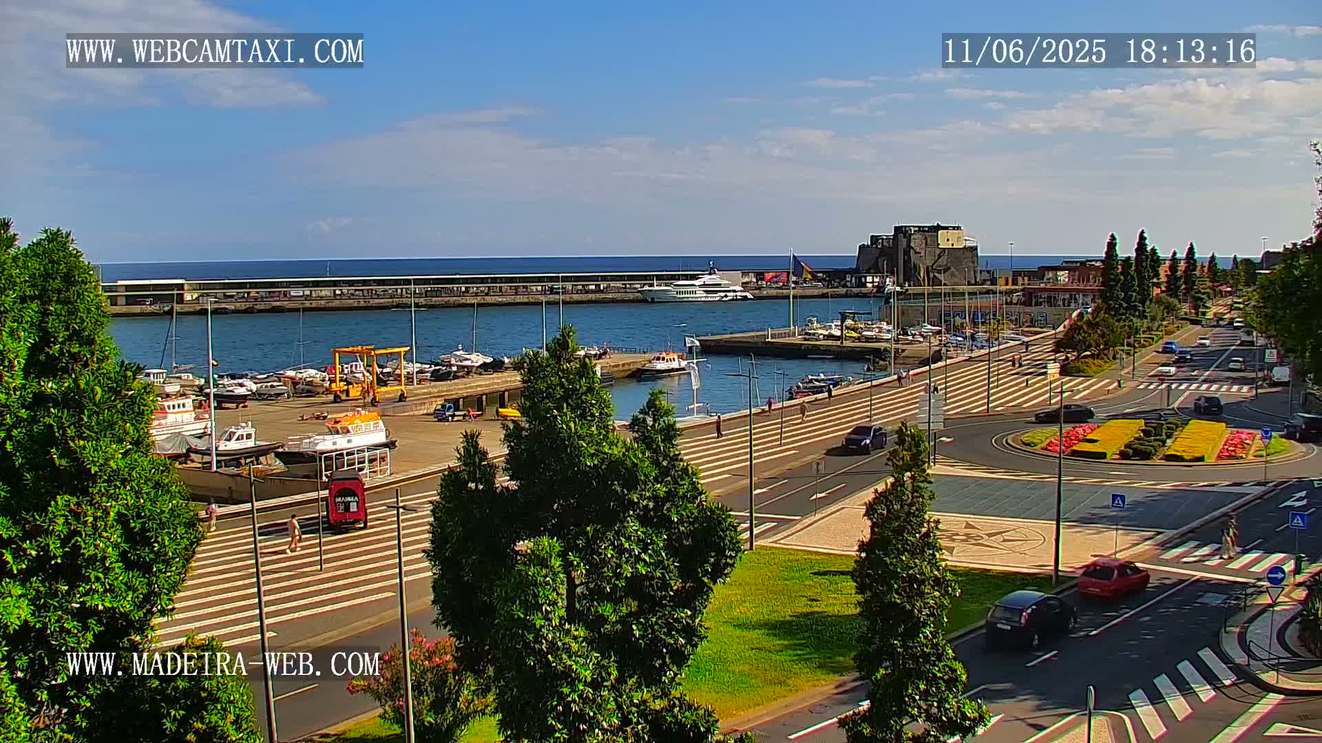A harbor filled with boats is seen from an elevated vantage point on a sunny day, with a paved area and roadway leading to a roundabout with landscaping.