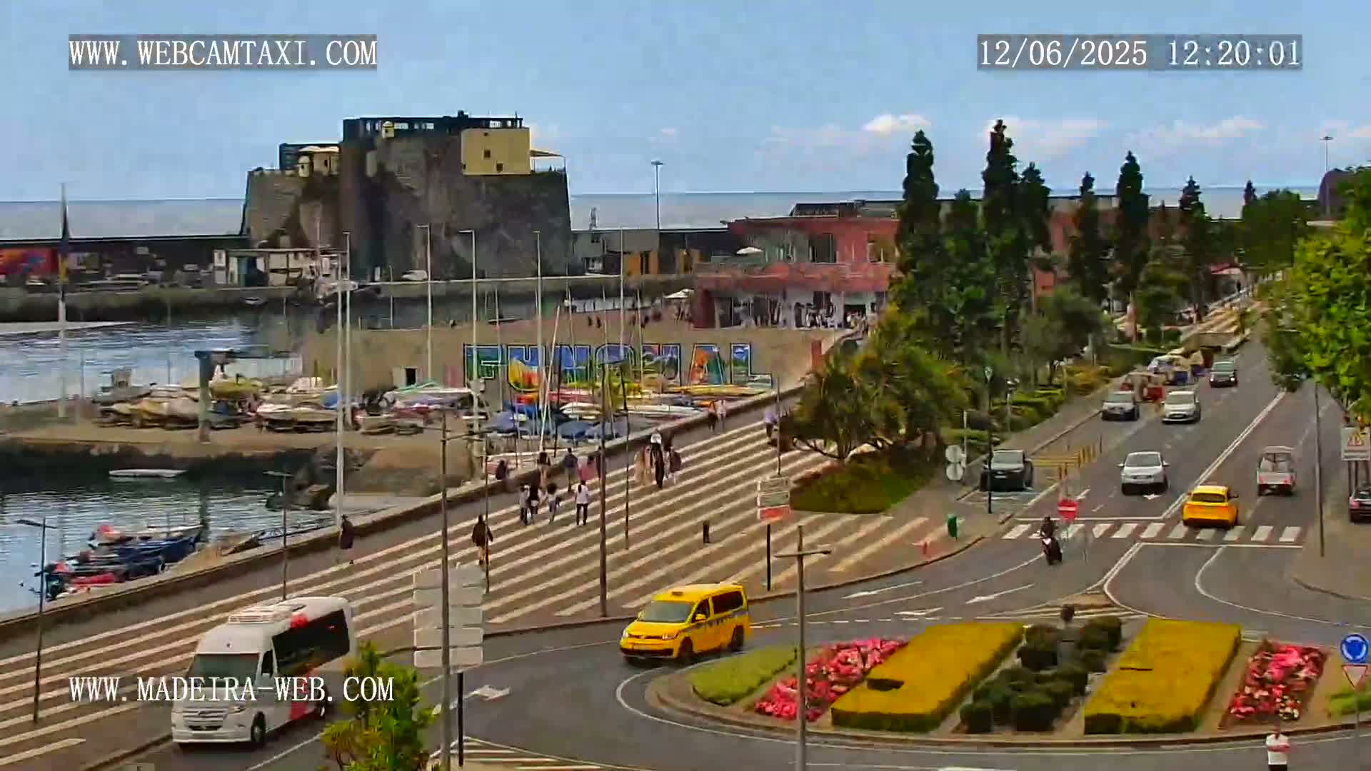 A harbor with docked boats, a stone fortress, and a roadway with cars and pedestrians is visible on a sunny day.