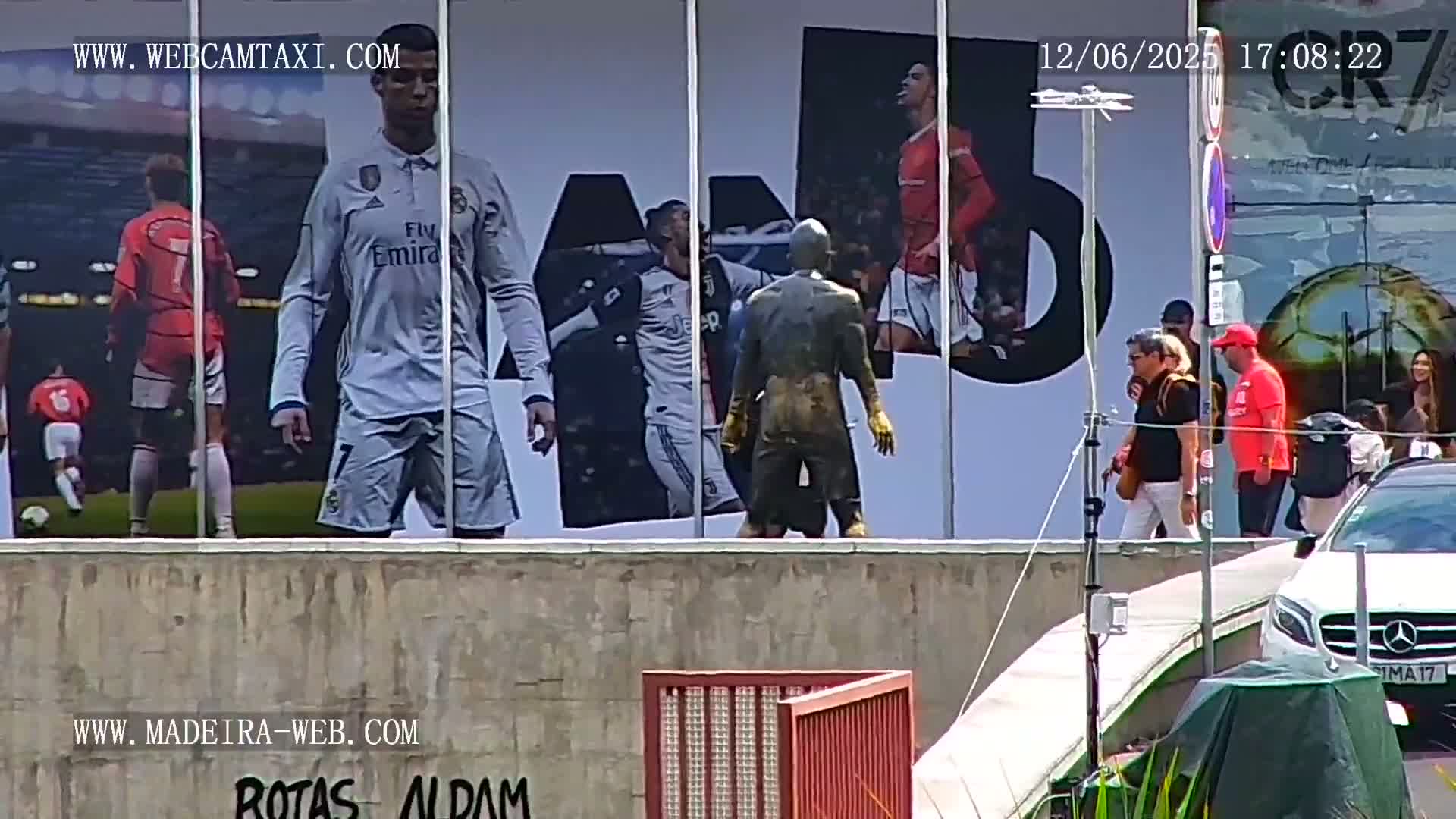 A large billboard displaying images of Cristiano Ronaldo in various soccer uniforms is visible behind several pedestrians walking on a sunny day.